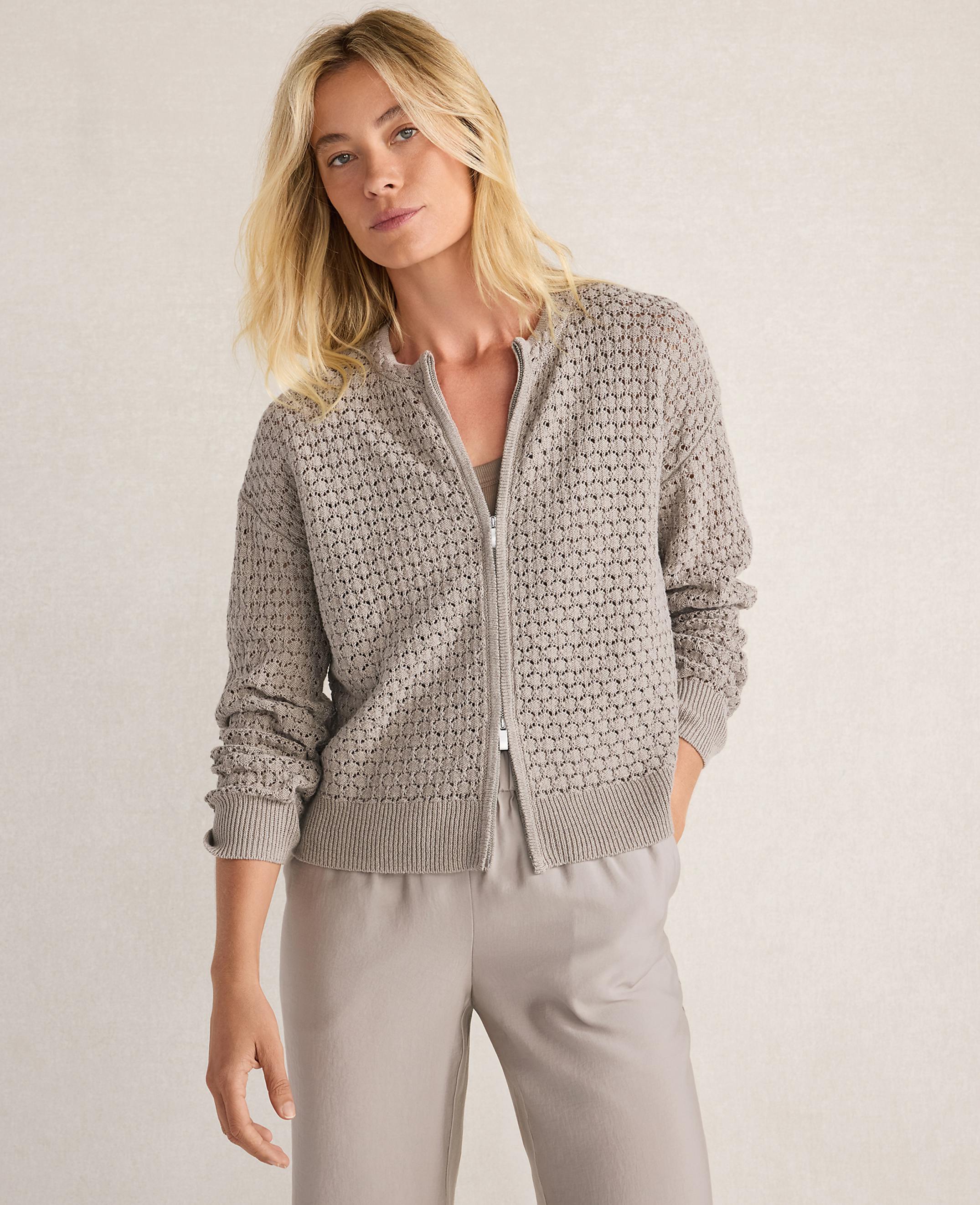 A blonde woman in a textured gray zip-up cardigan and light gray pants stands against a neutral, textured background.
