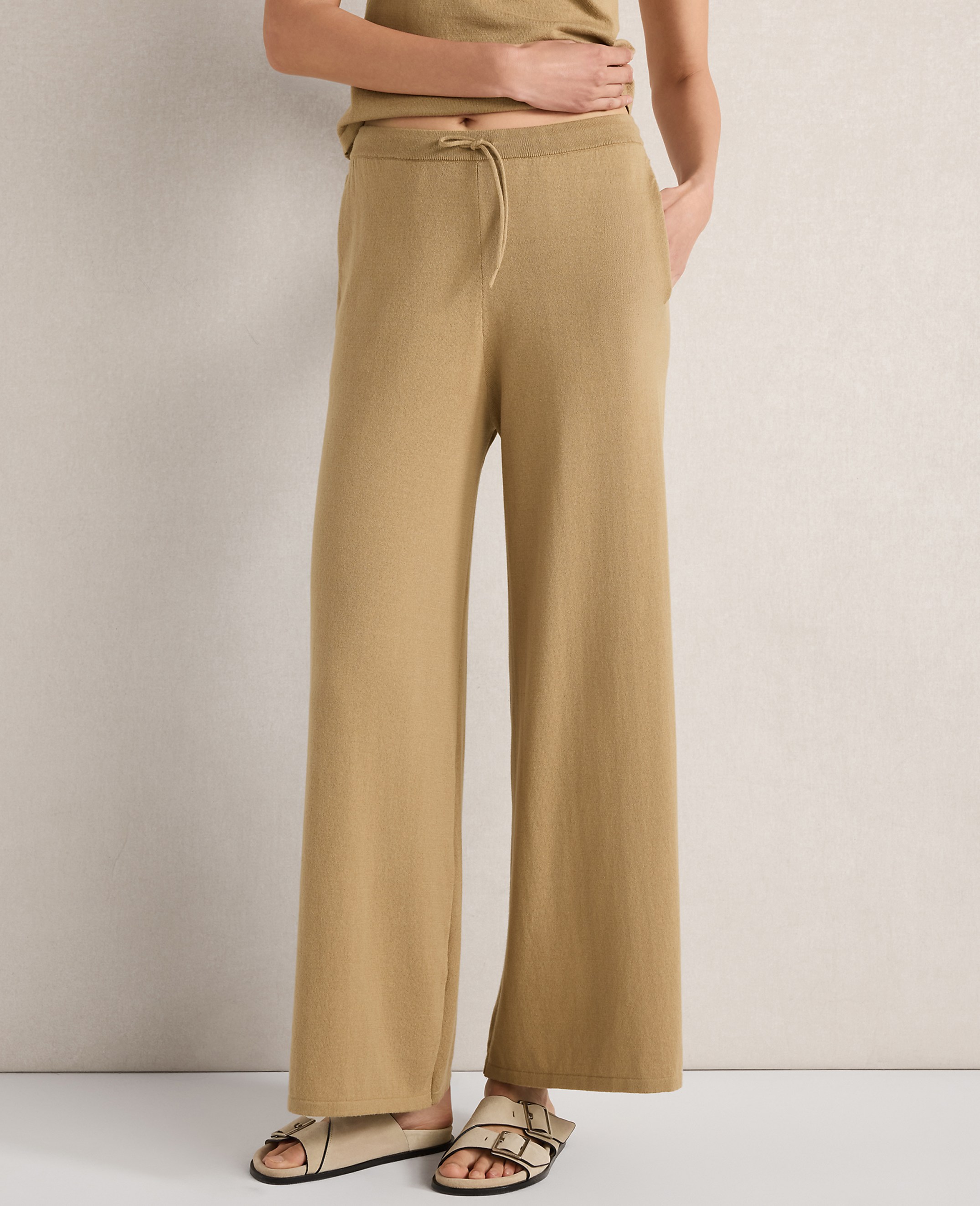 A person stands in tan wide-leg pants and sandals against a neutral, textured wall.