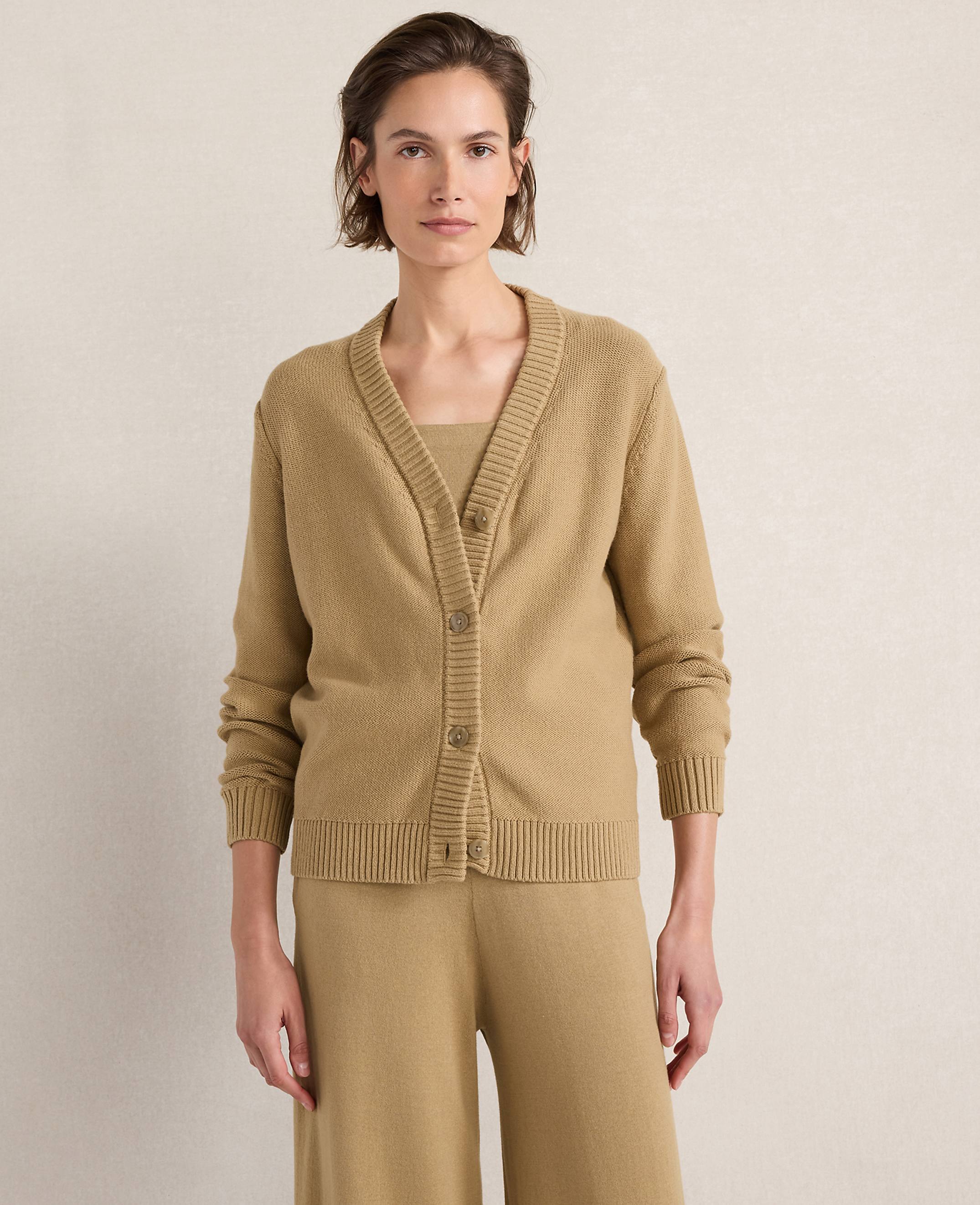 A woman in a tan cardigan and matching pants stands against a plain, light-colored background.