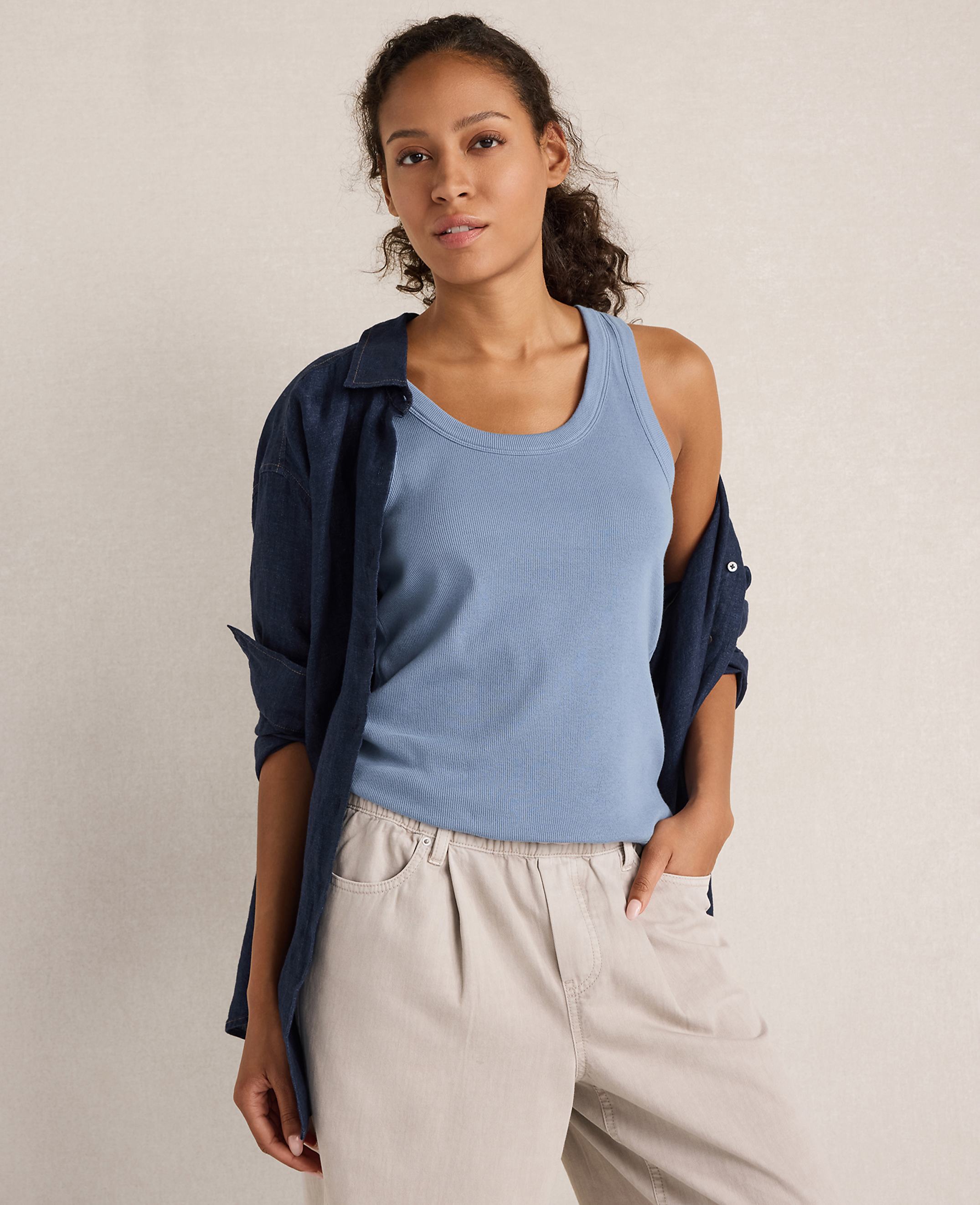 A woman in a blue tank top and tan pants stands against a plain, light-colored background.