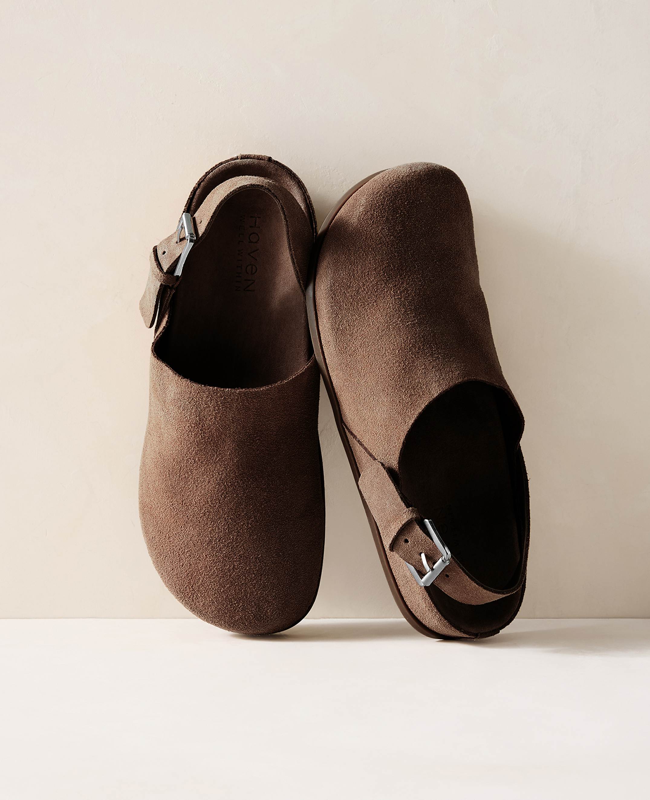 A pair of brown suede clogs with buckles rests on a light-colored surface against a soft, neutral background.
