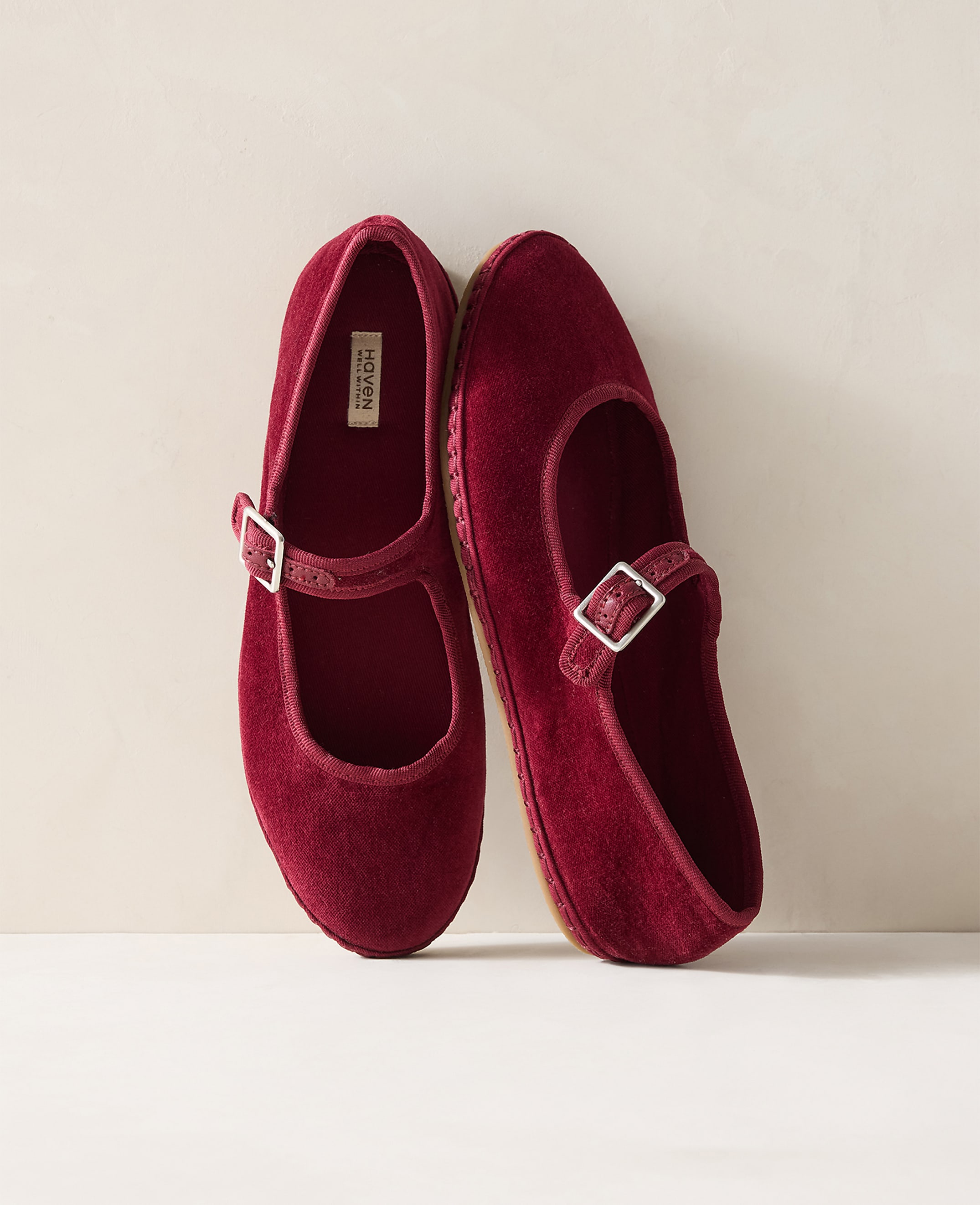 A pair of burgundy velvet Mary Jane slippers with buckle straps rest on a white surface against a plain, light-colored background.