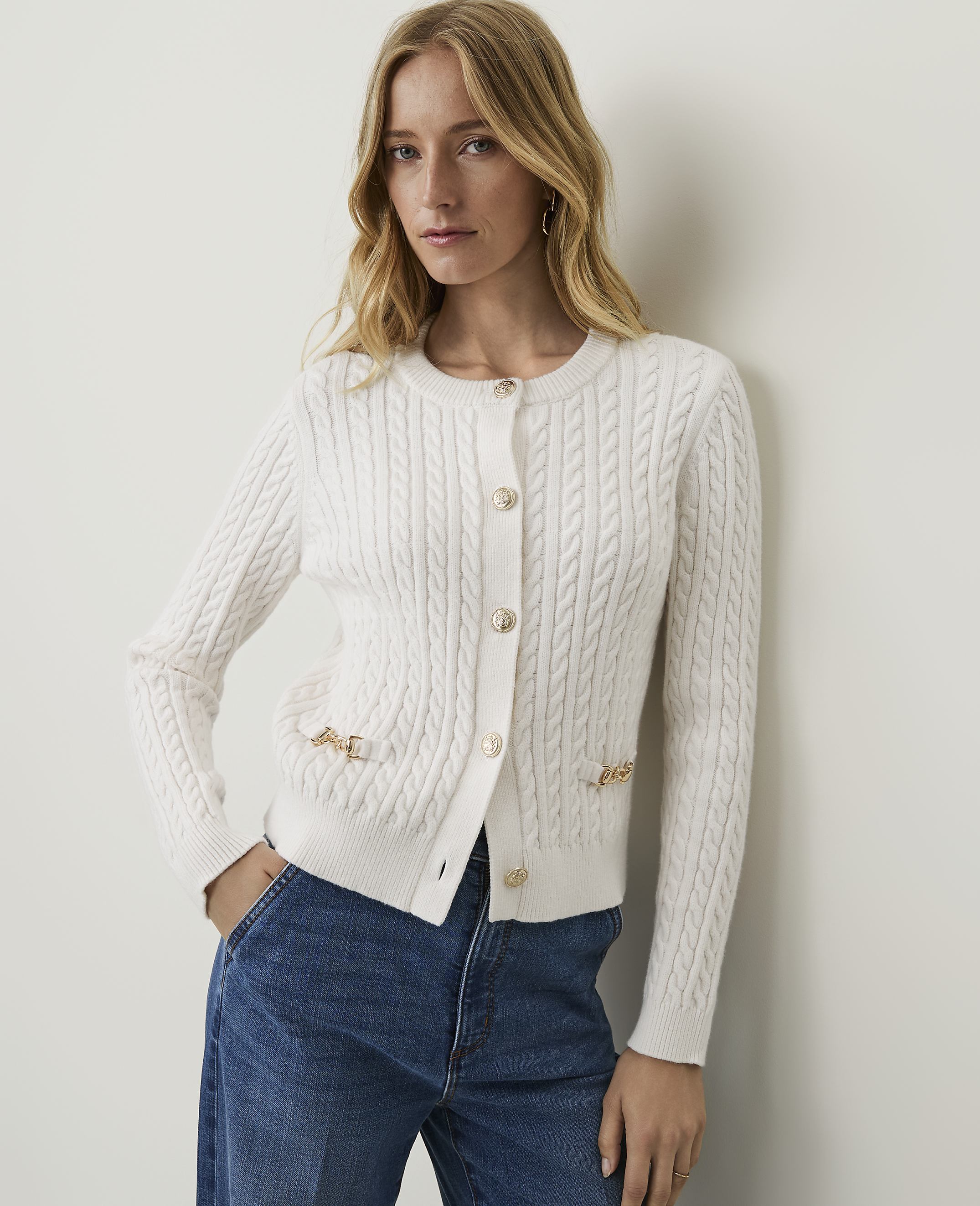 A woman in a white cable-knit cardigan and blue jeans stands against a plain white background.