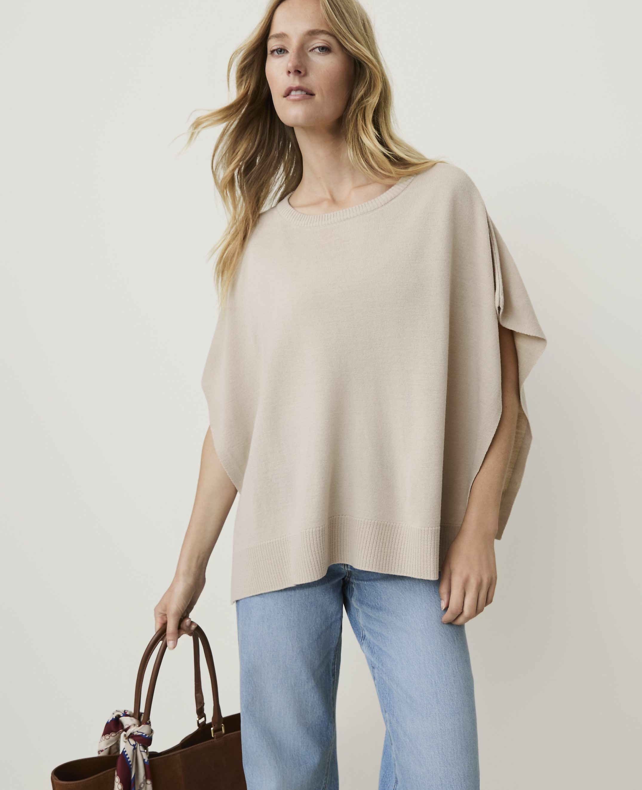 A woman in a beige poncho and light blue jeans holds a brown tote bag with a scarf in front of a plain beige background.