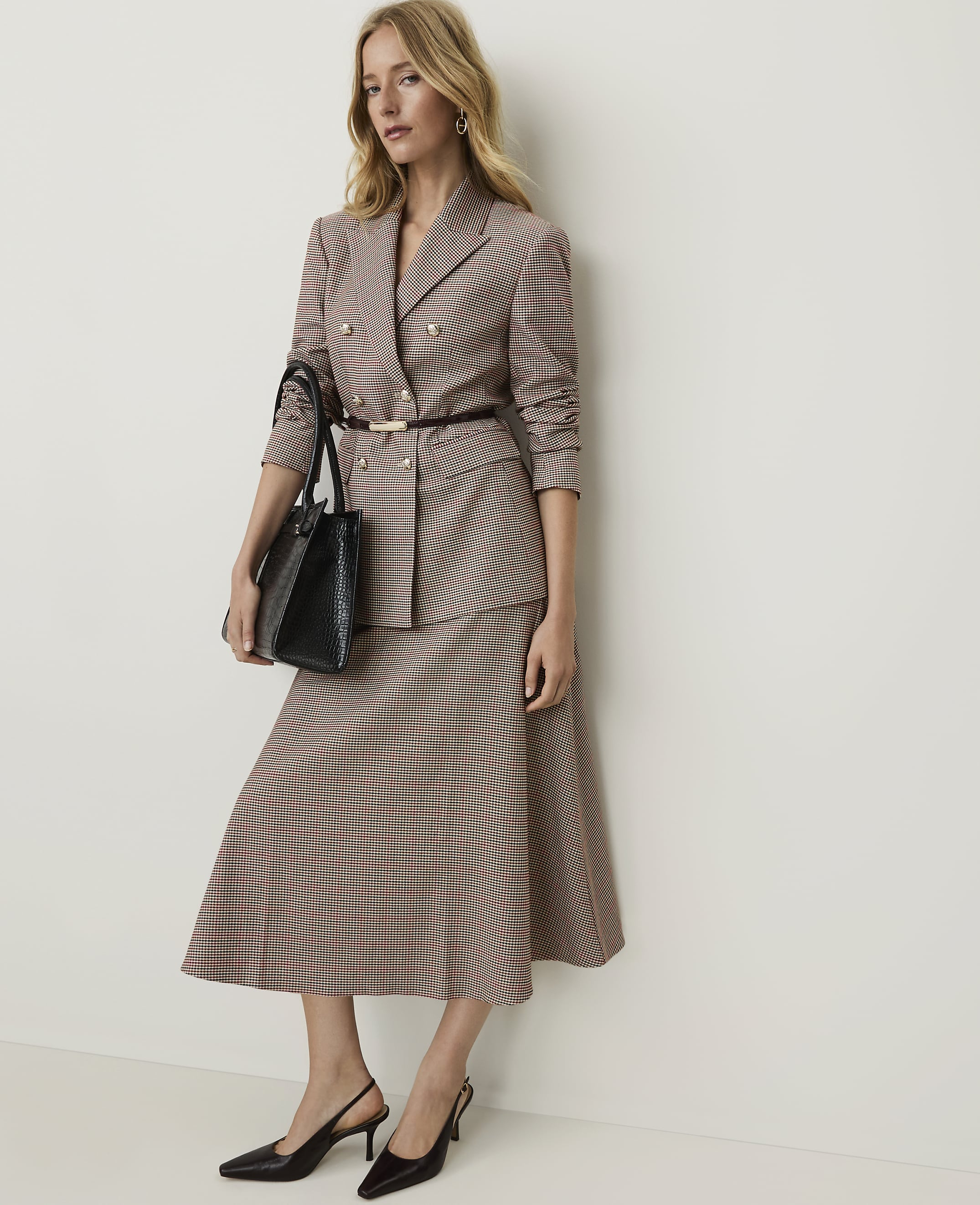 A woman in a patterned suit and heels stands holding a black handbag against a plain, light-colored wall.