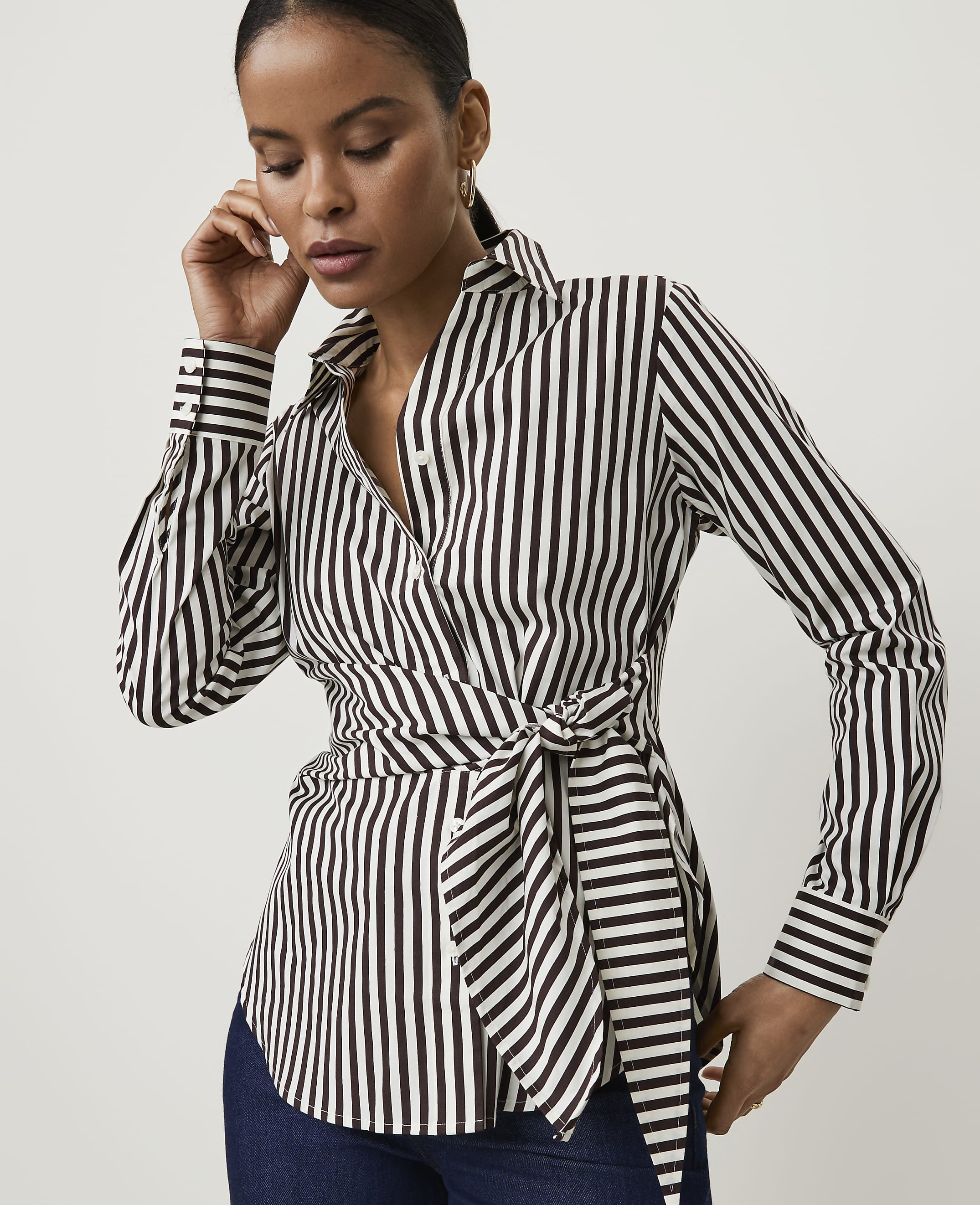 A woman in a black and white striped wrap shirt with a tie at the waist poses against a plain white background.