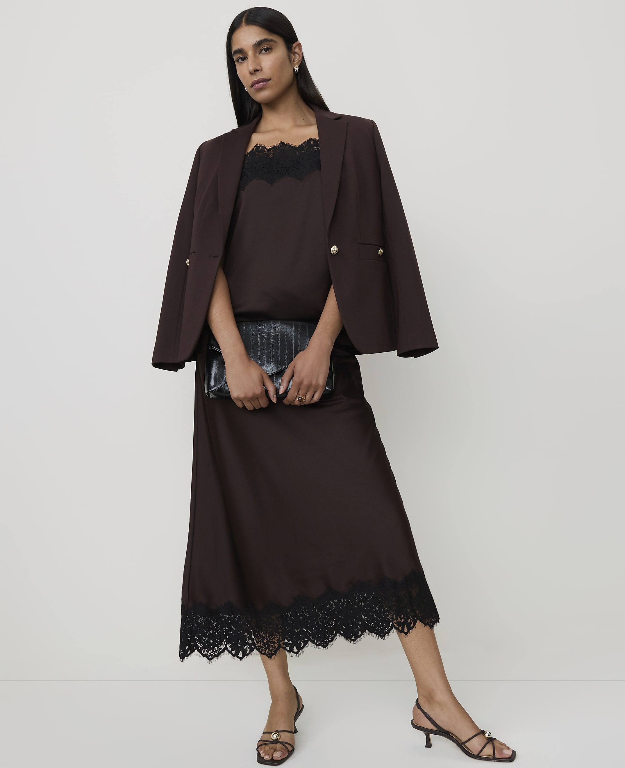 A woman in a brown slip dress and blazer holds a black clutch in front of a plain white background.