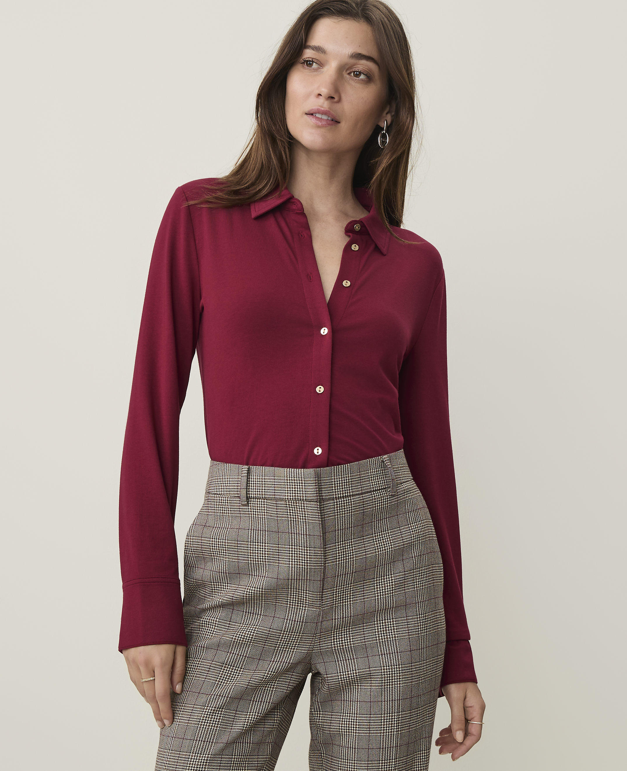 A woman in a maroon button-up shirt and plaid pants stands against a plain, light-colored background.