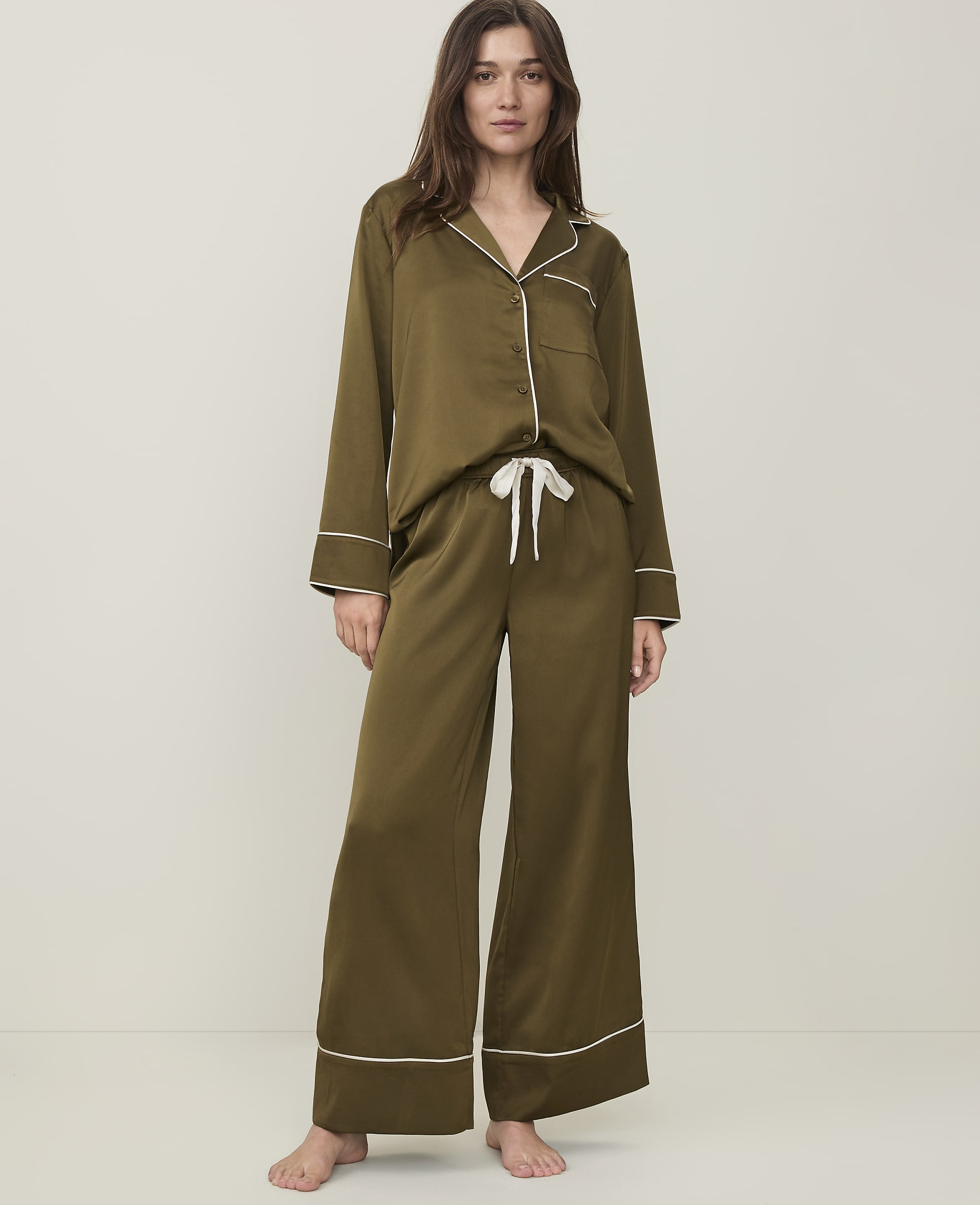A woman stands in olive green satin pajamas with white piping against a plain, light-colored background.