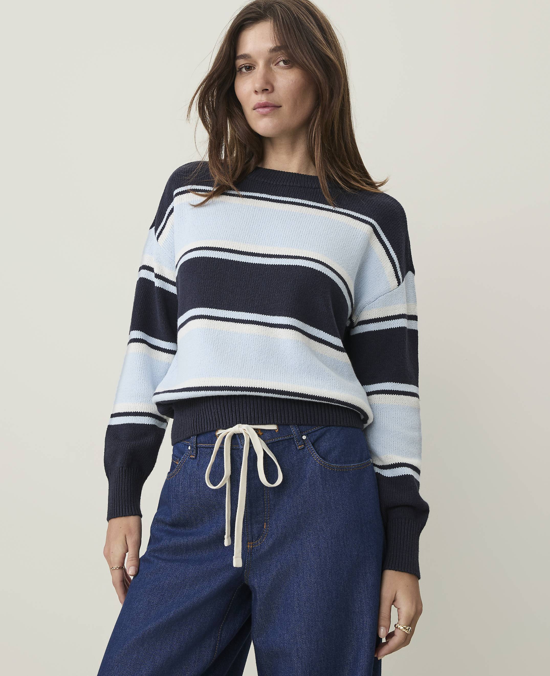 A woman in a striped sweater and jeans stands against a plain, light background.