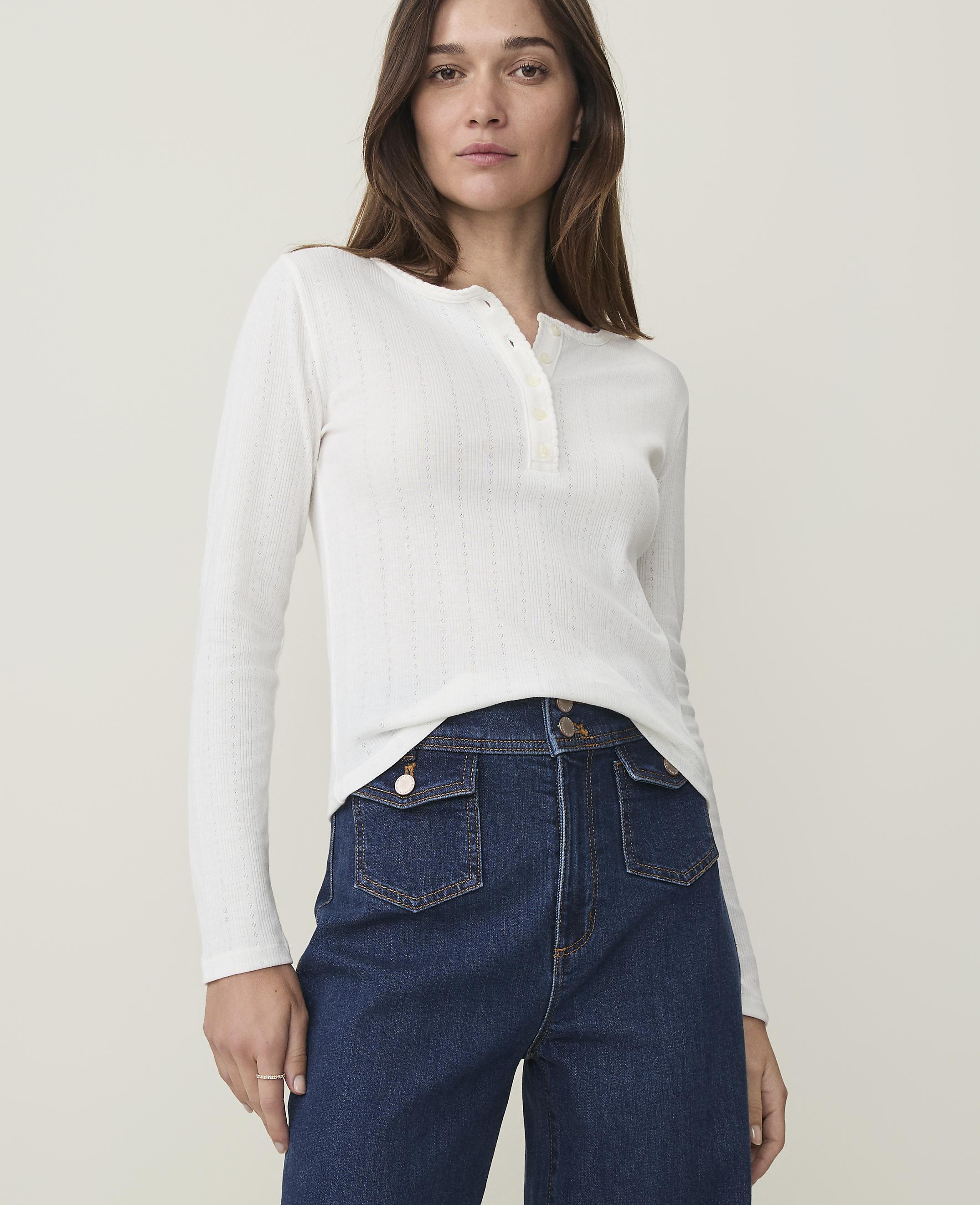 A woman in a white henley shirt and dark wash jeans stands against a plain, light background.
