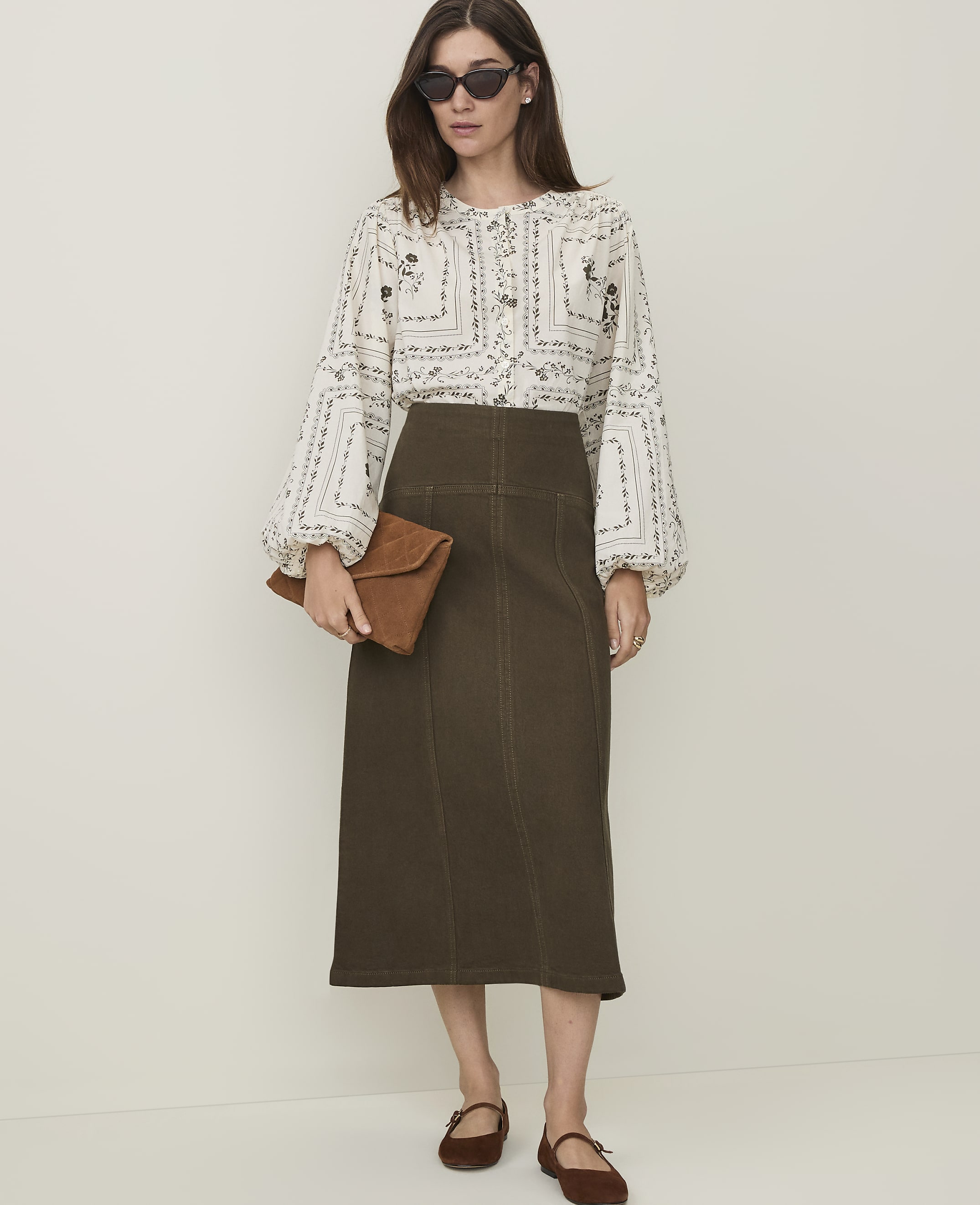 A woman in a patterned blouse and olive green skirt stands against a plain, light-colored background, holding a brown clutch.
