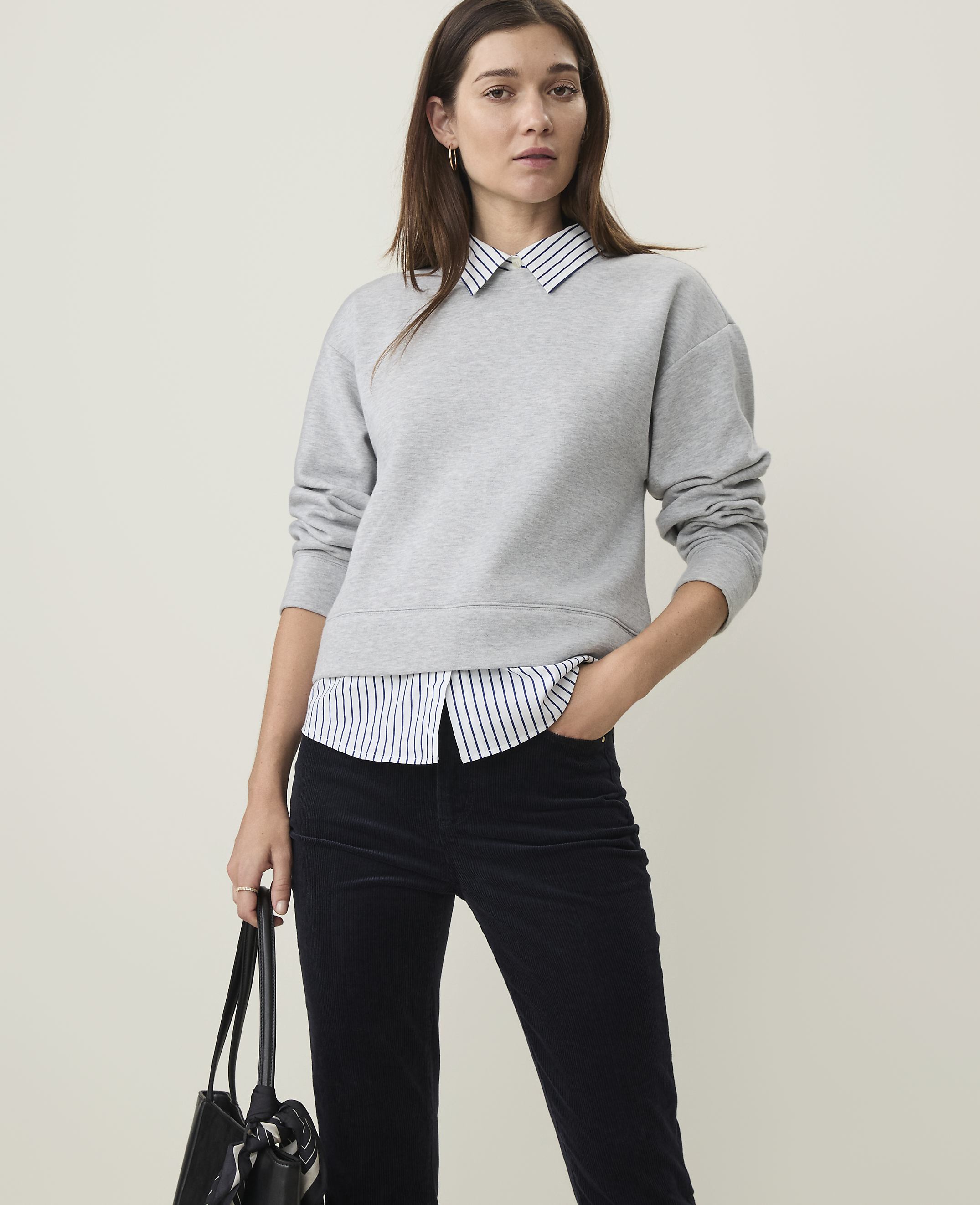 A woman in a grey sweatshirt layered over a striped collared shirt and dark pants stands against a plain, light background.