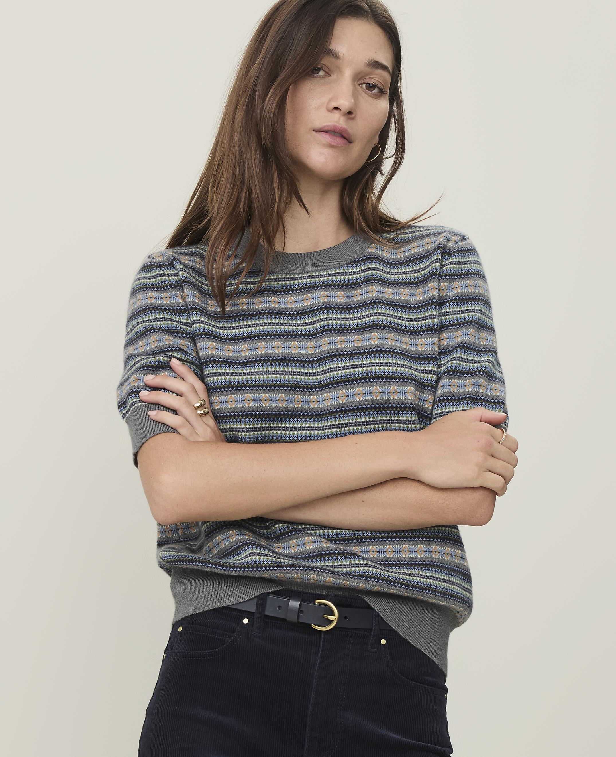 A woman with crossed arms in a patterned sweater stands against a plain, light-colored background.