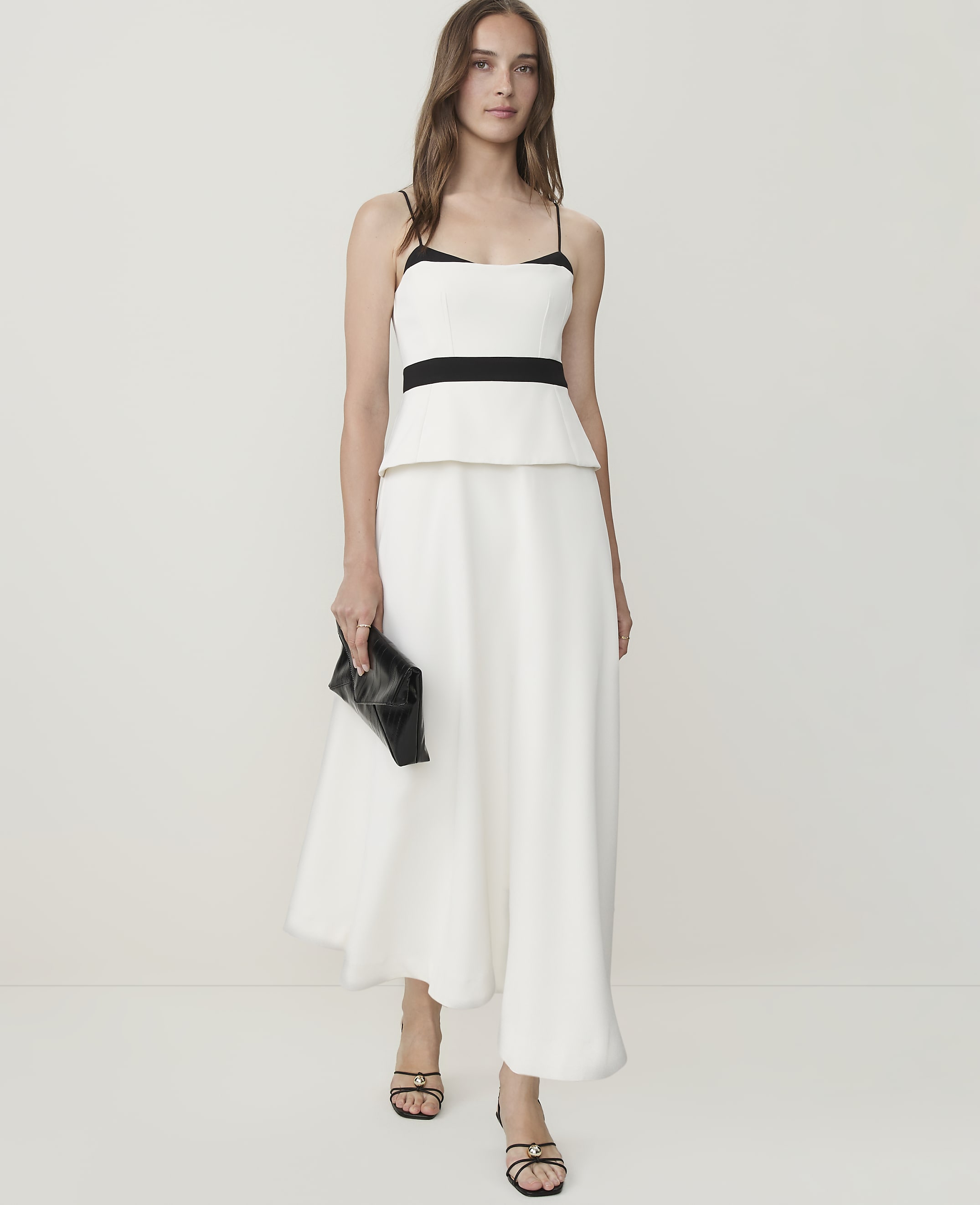A woman in a white peplum top and wide-leg pants holds a black clutch against a plain white background.
