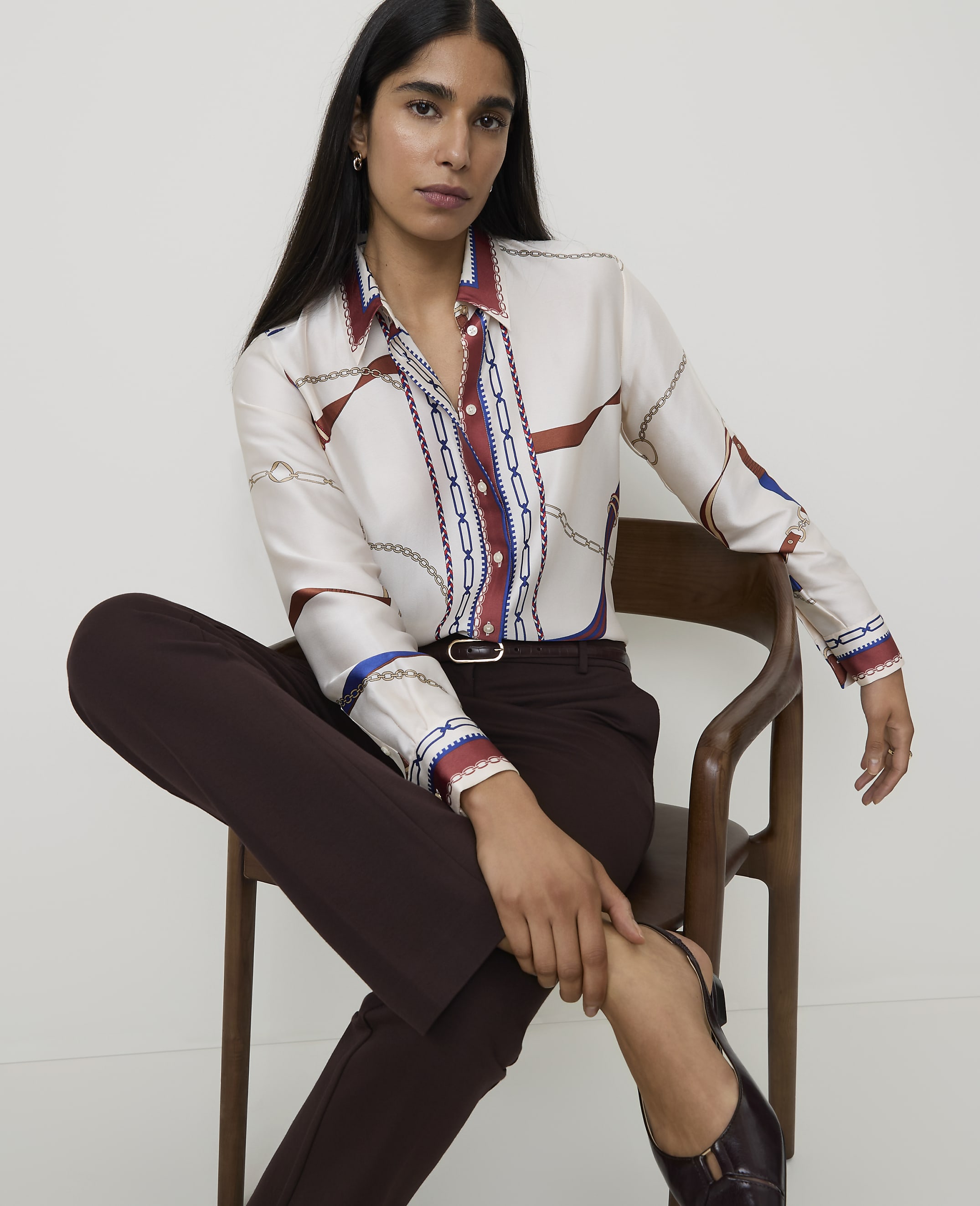 A woman in a patterned silk shirt and dark trousers sits on a wooden chair against a plain background.