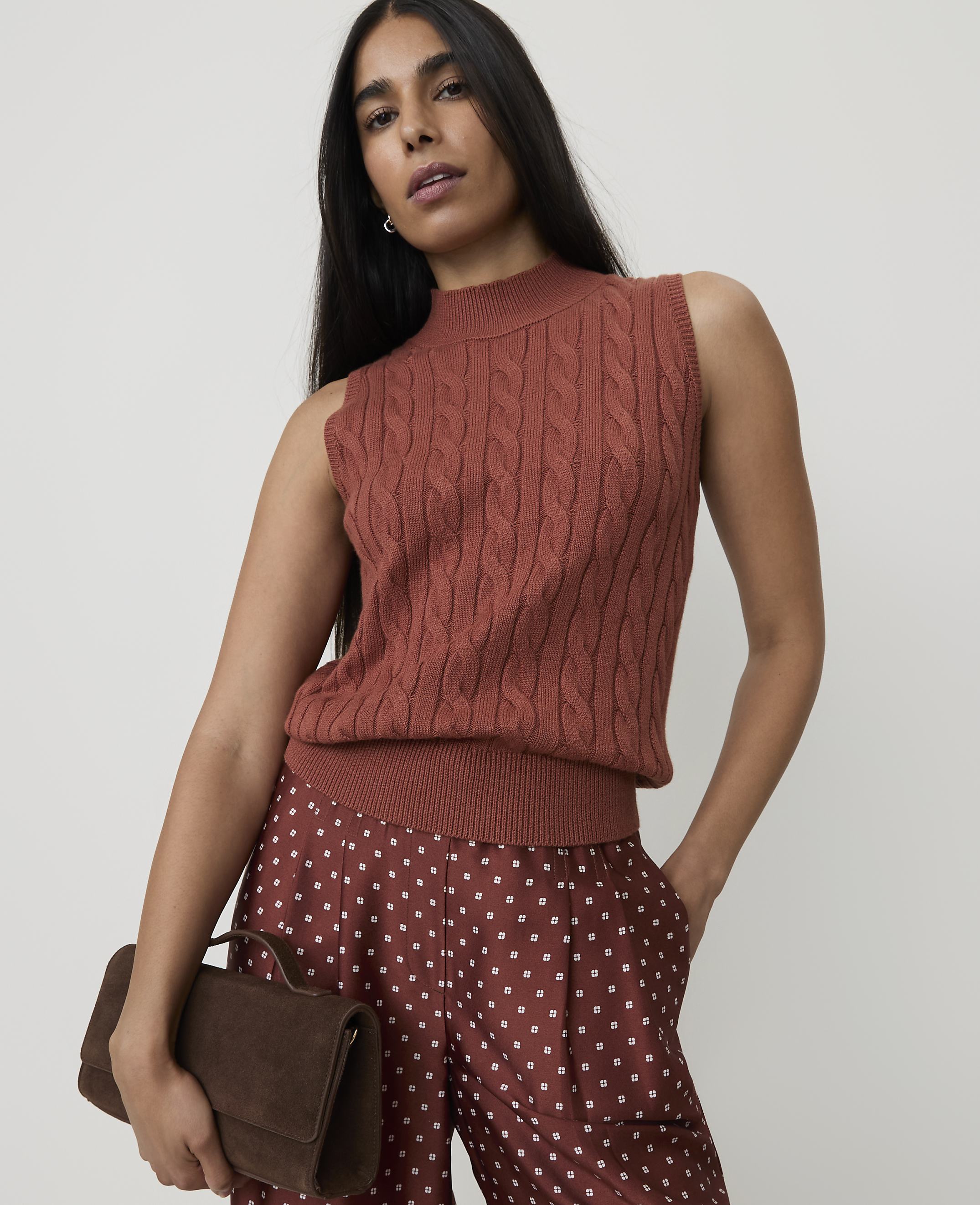 A woman in a rust-colored cable-knit sleeveless turtleneck and polka-dot pants holds a brown clutch against a plain background.