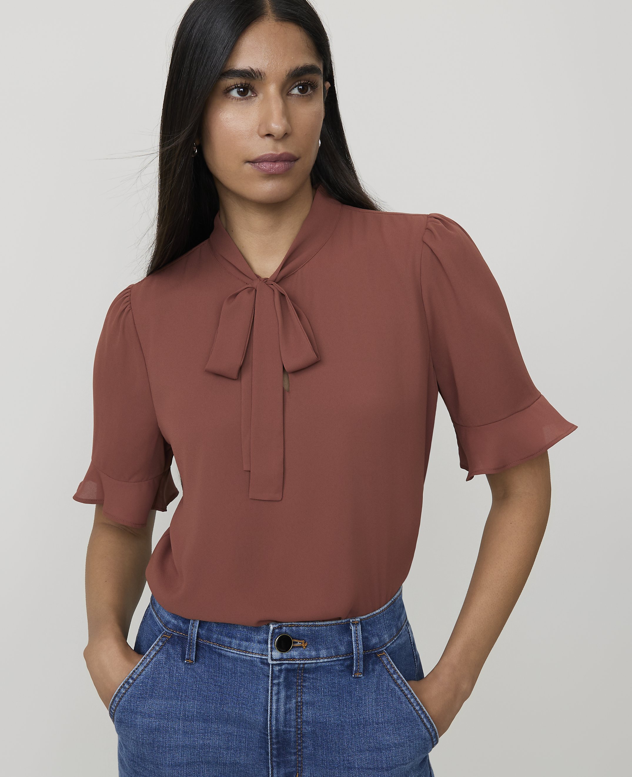 A woman in a rust-colored bow-neck blouse and blue jeans stands against a plain white background.