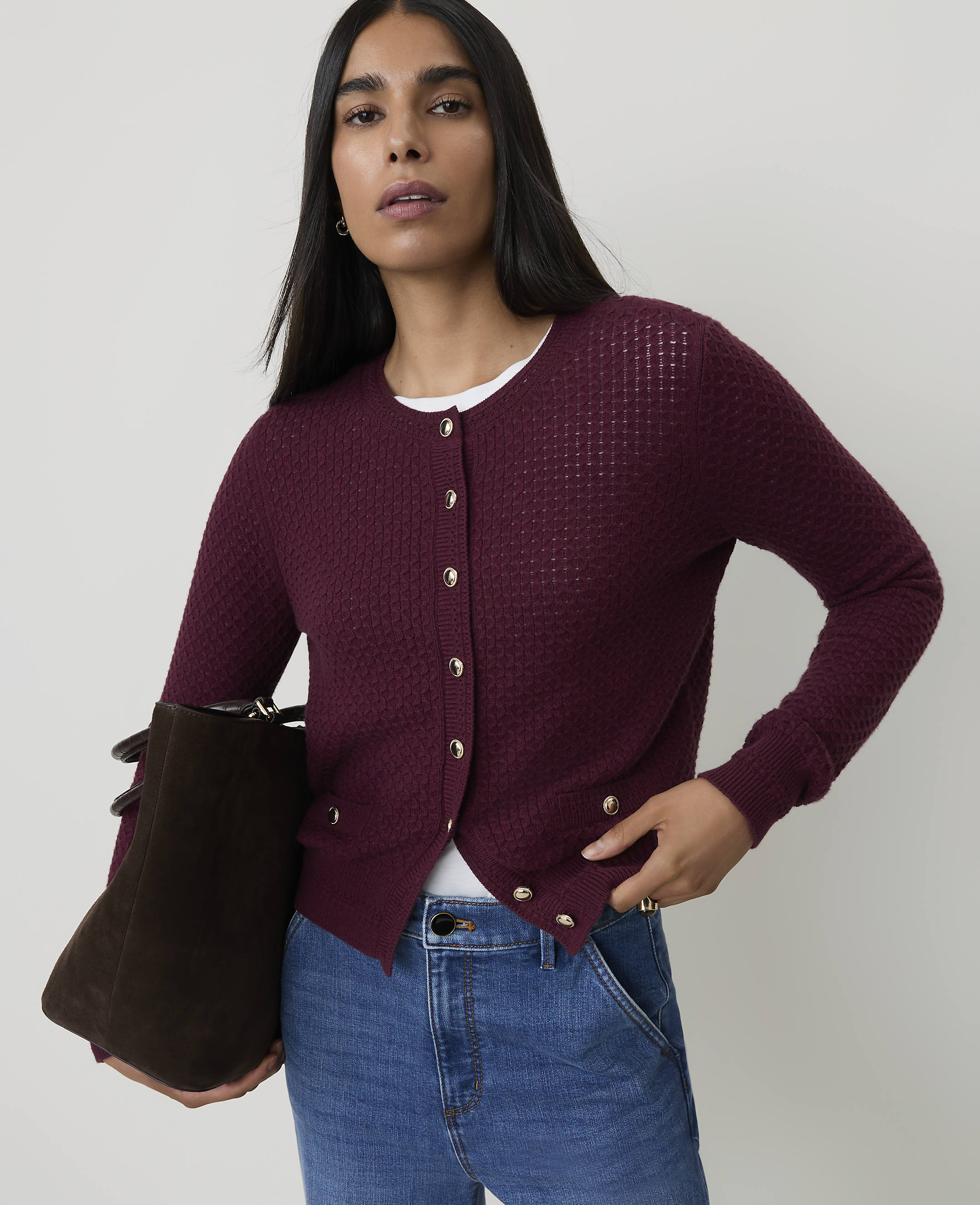 A woman in a burgundy cardigan and jeans stands against a plain background, holding a dark brown bag.