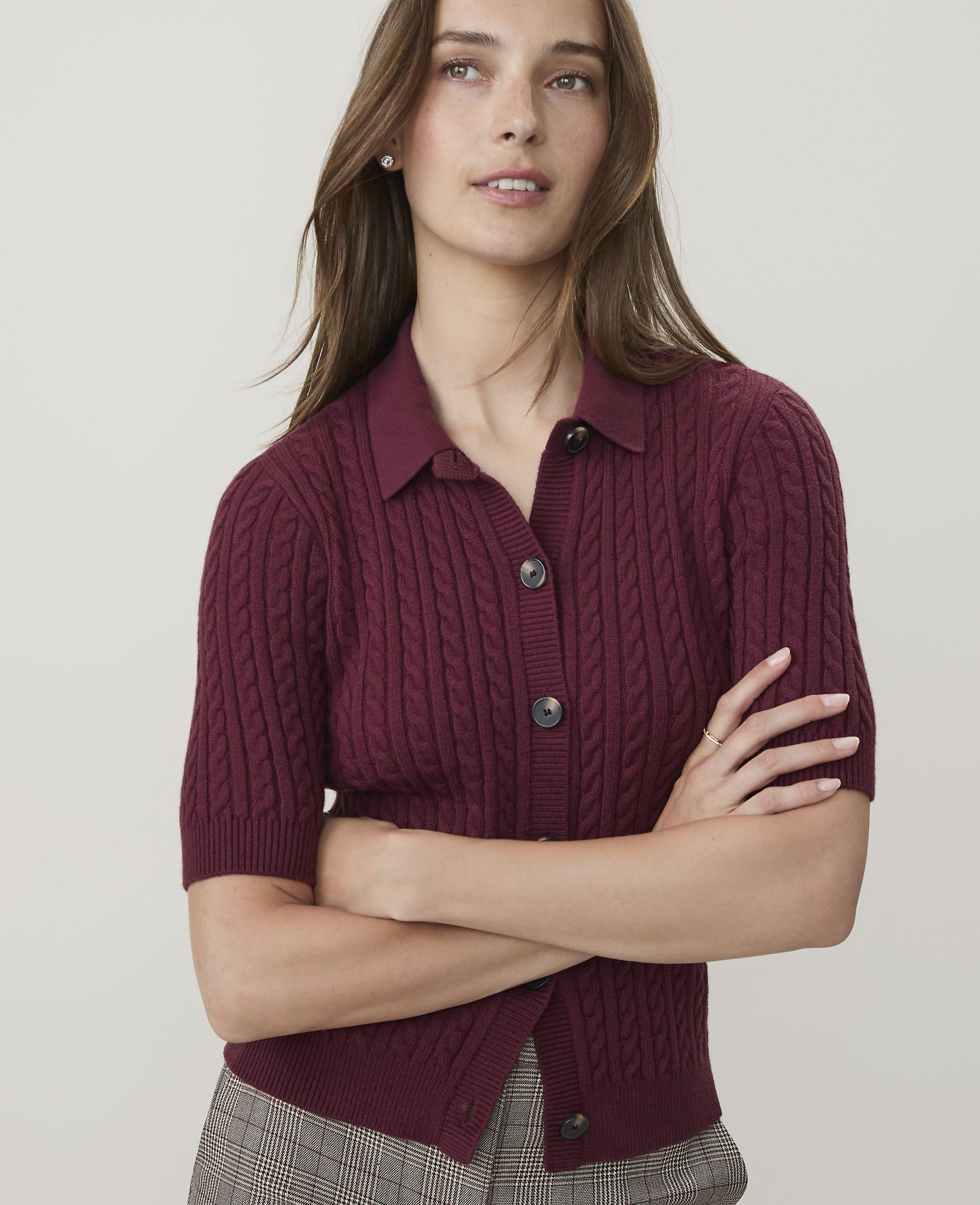 A woman in a burgundy cable-knit polo shirt with her arms crossed stands against a plain white background.