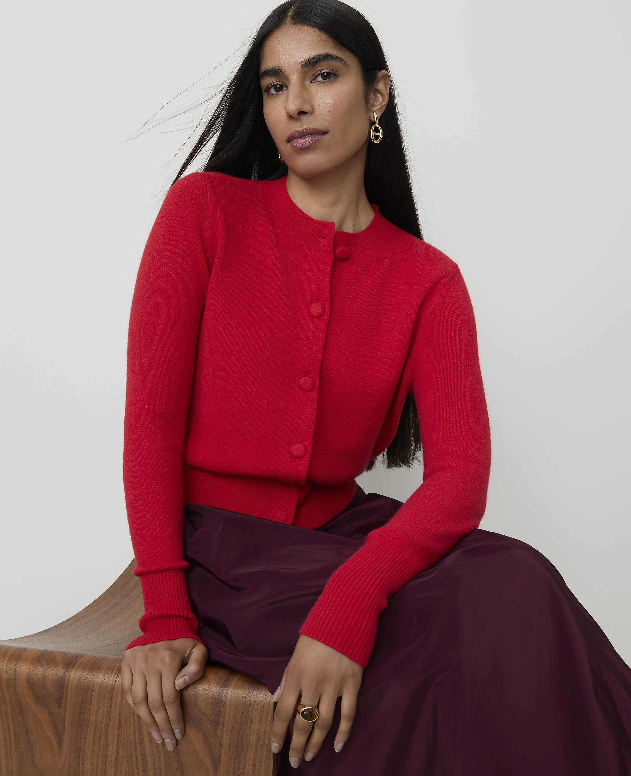 A woman in a red cardigan and plum skirt sits on a wooden stool against a plain background.