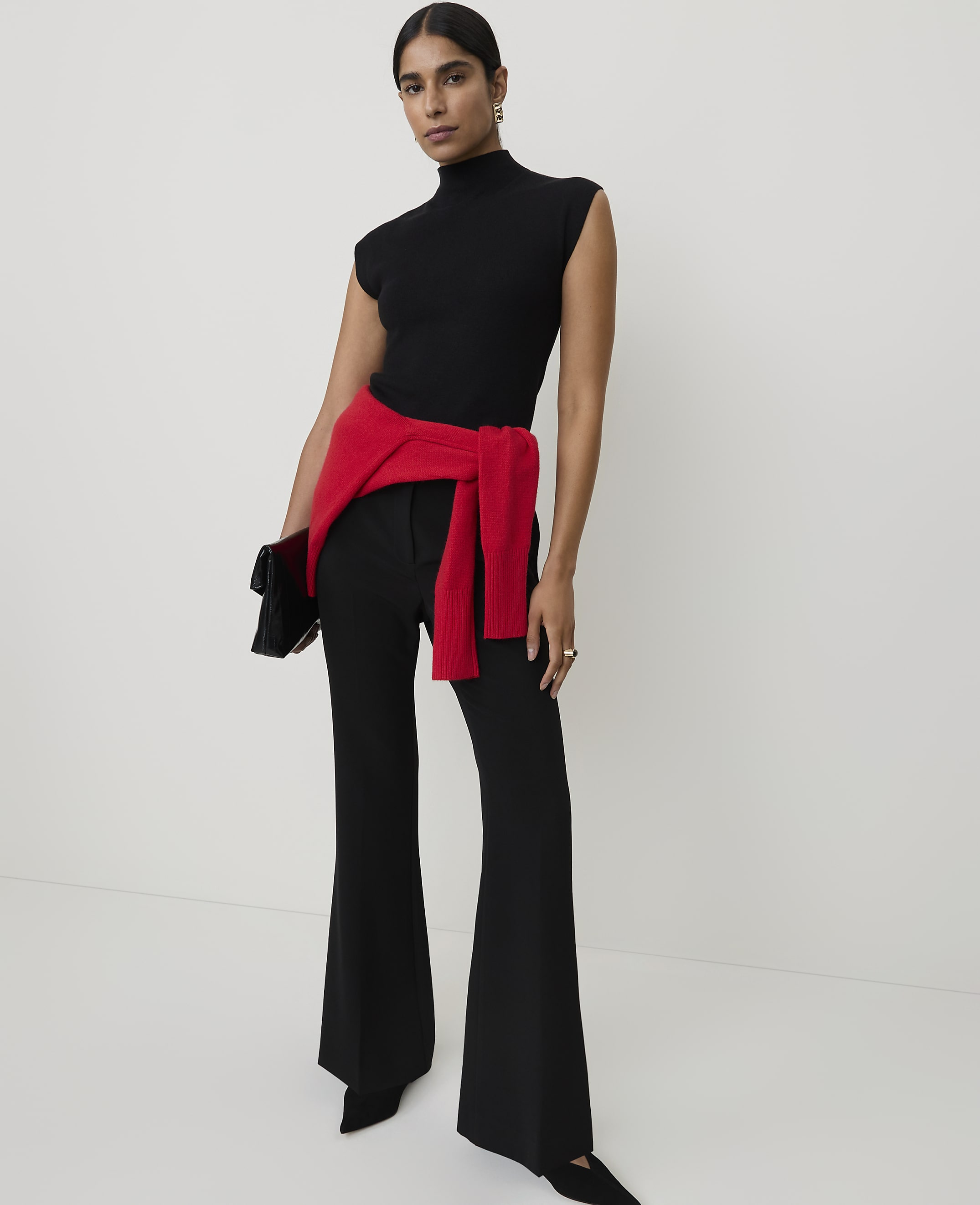 A woman in a black turtleneck and flared pants, with a red sweater tied around her waist, stands against a plain white background.