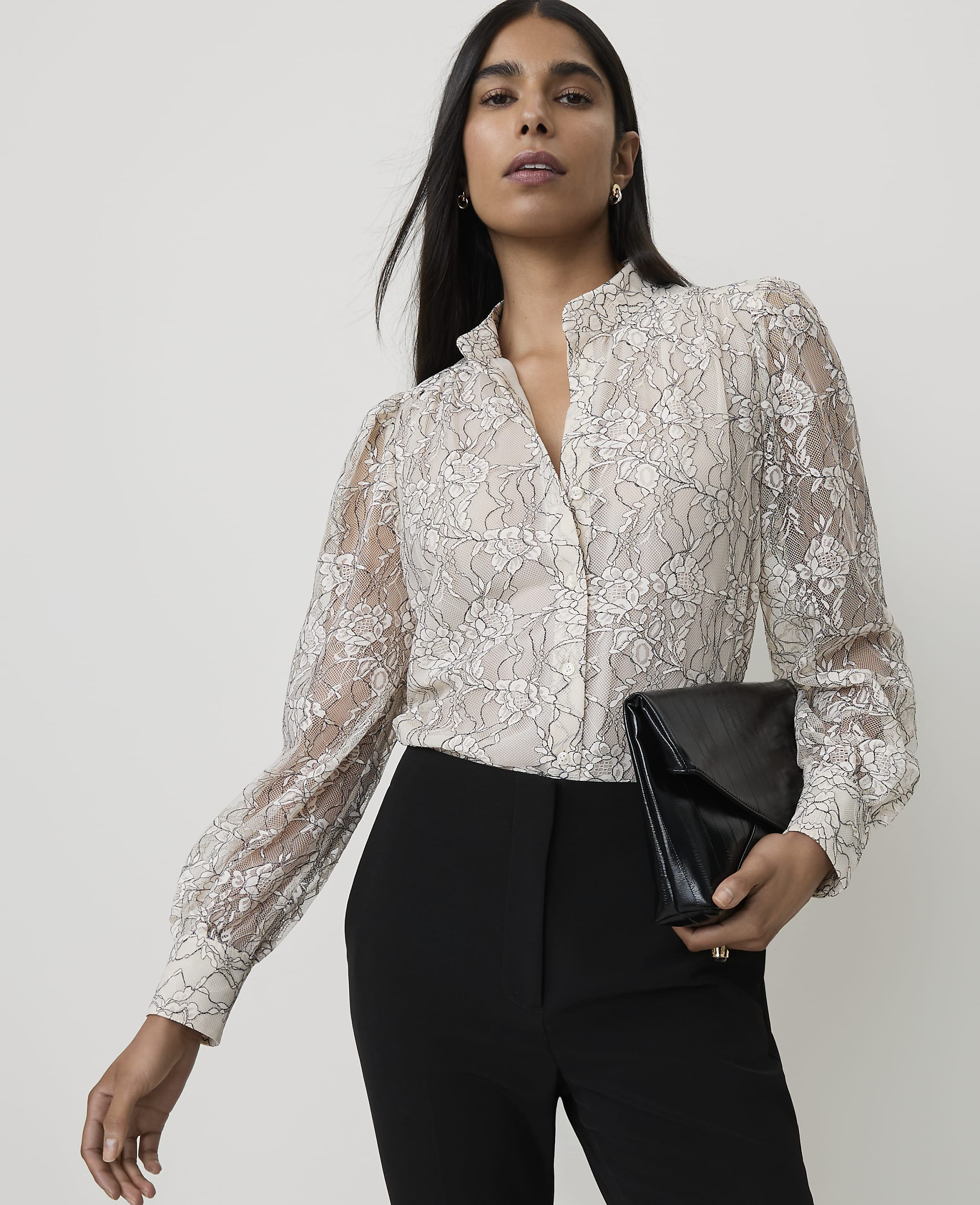 A woman in a lace blouse and black pants holds a black clutch against a plain background.