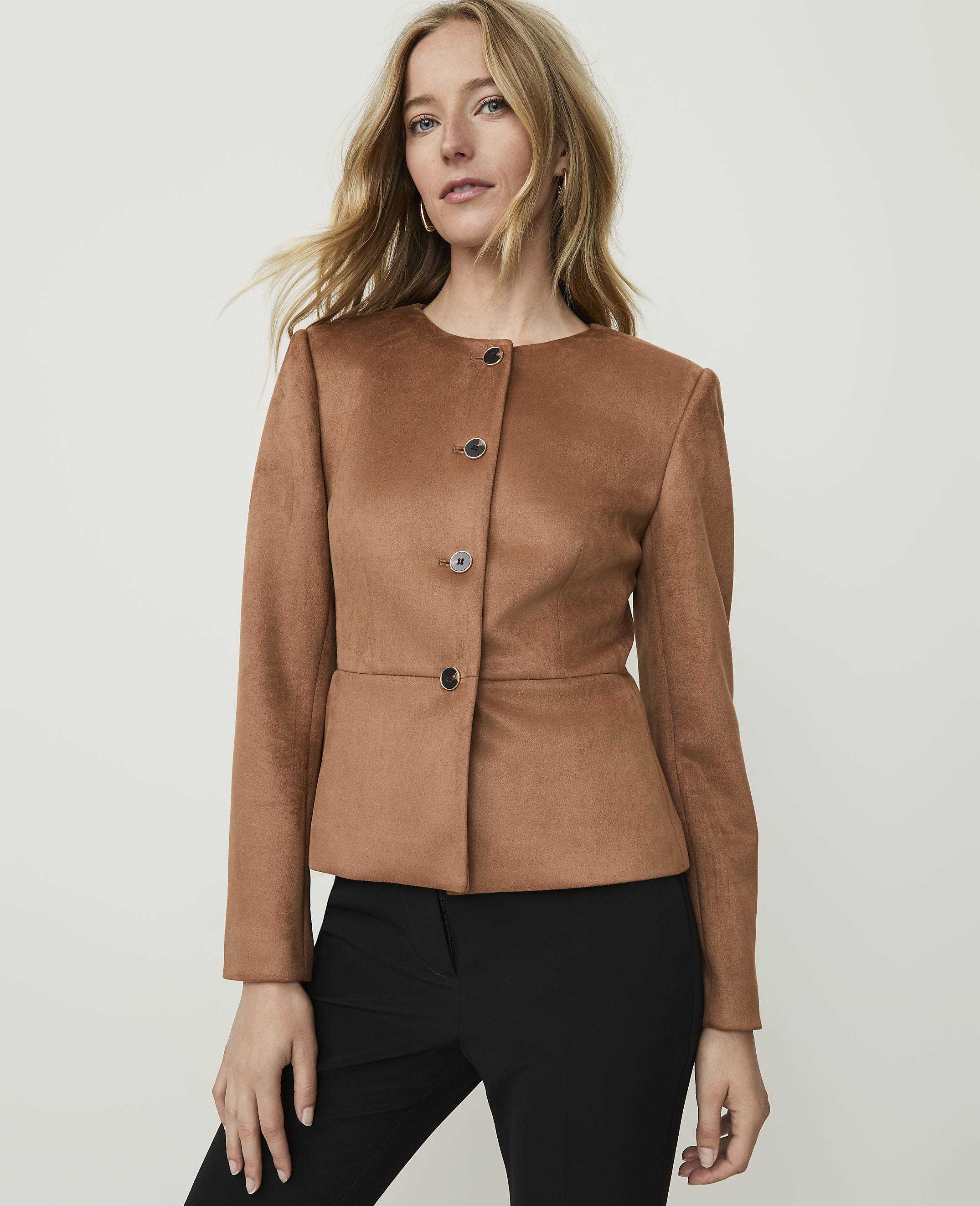 A blonde woman in a brown jacket and black pants stands against a plain, light gray background.