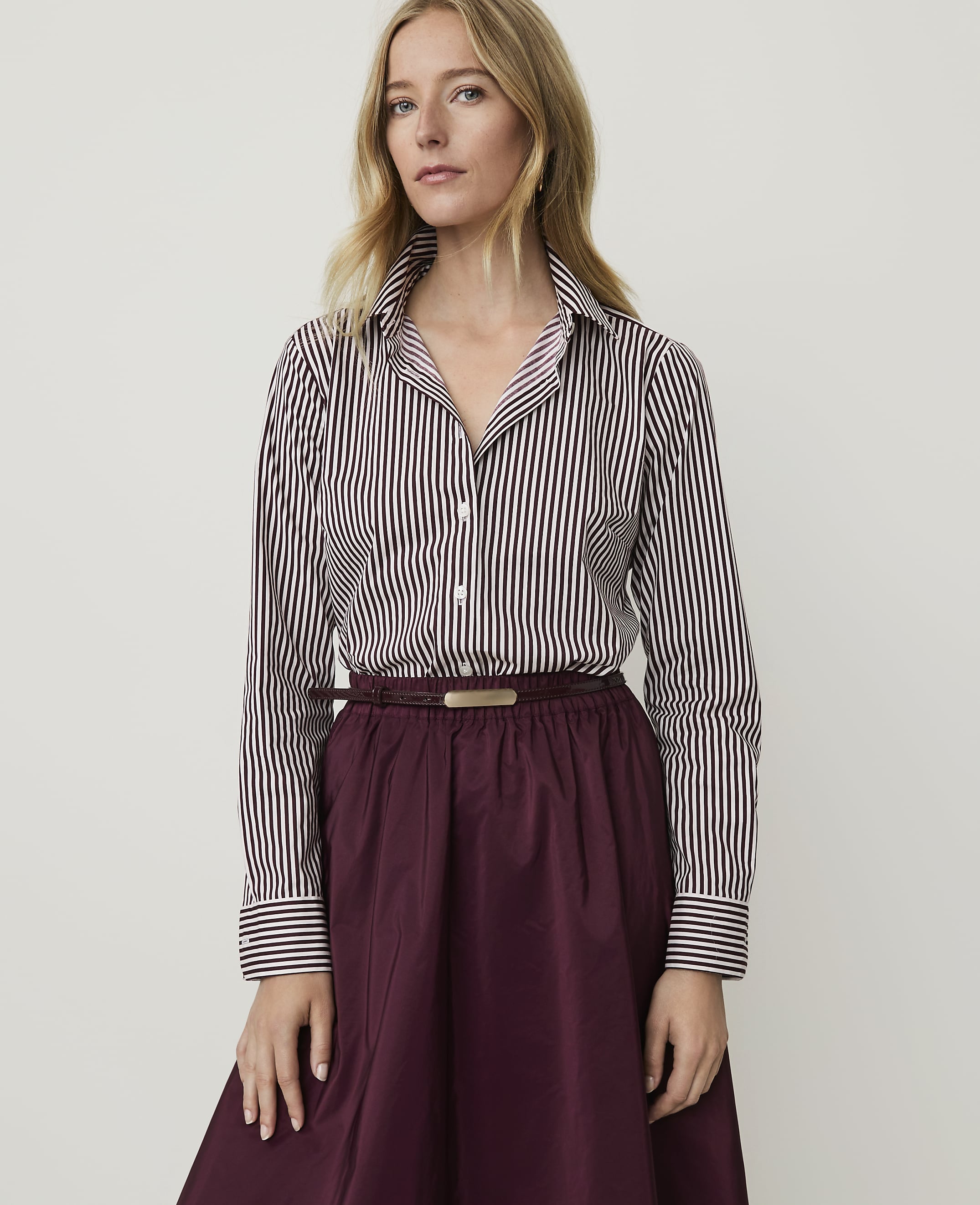 A woman in a striped shirt and burgundy skirt stands against a plain white background.