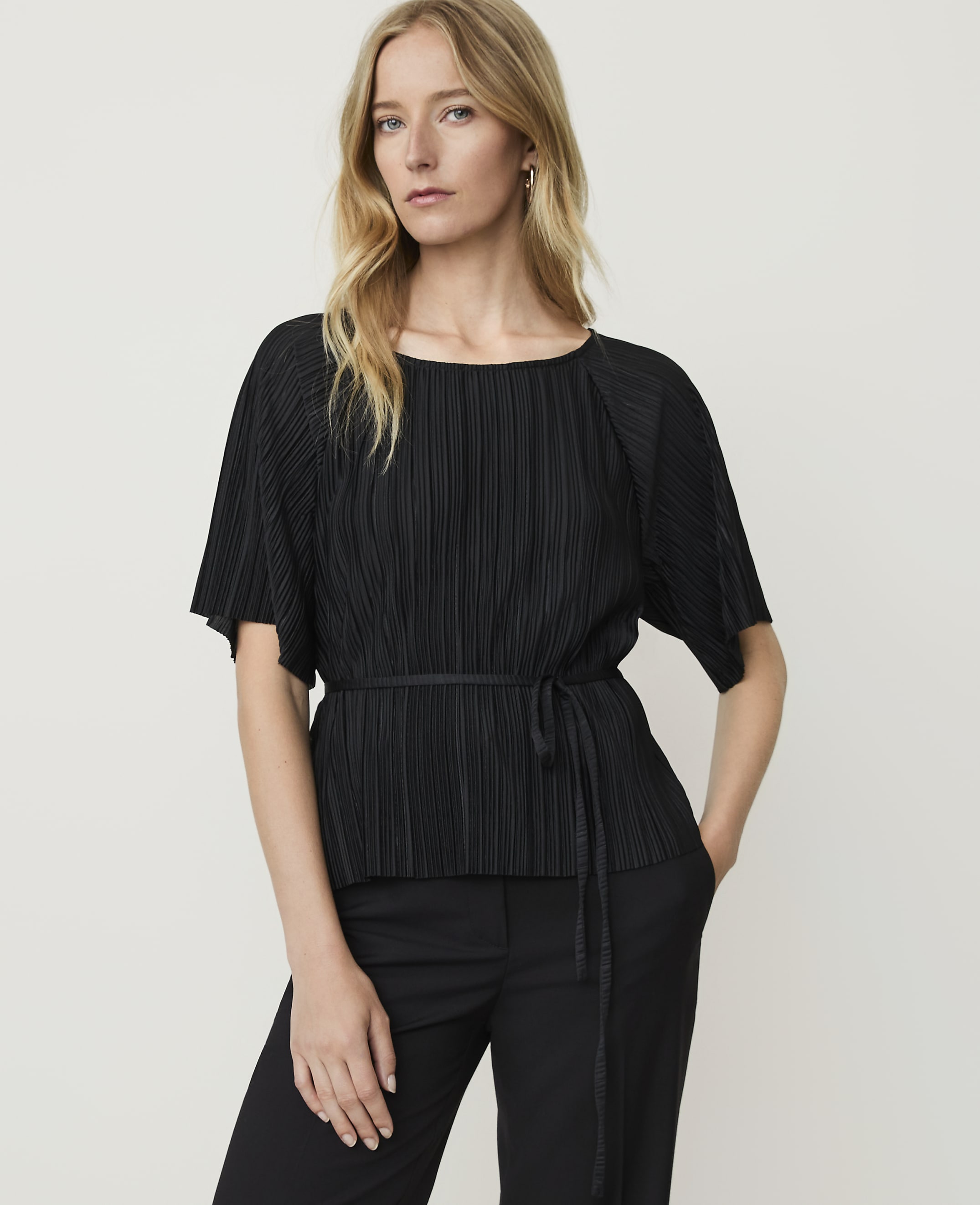 A blonde woman in a black pleated top and pants stands against a plain white background.