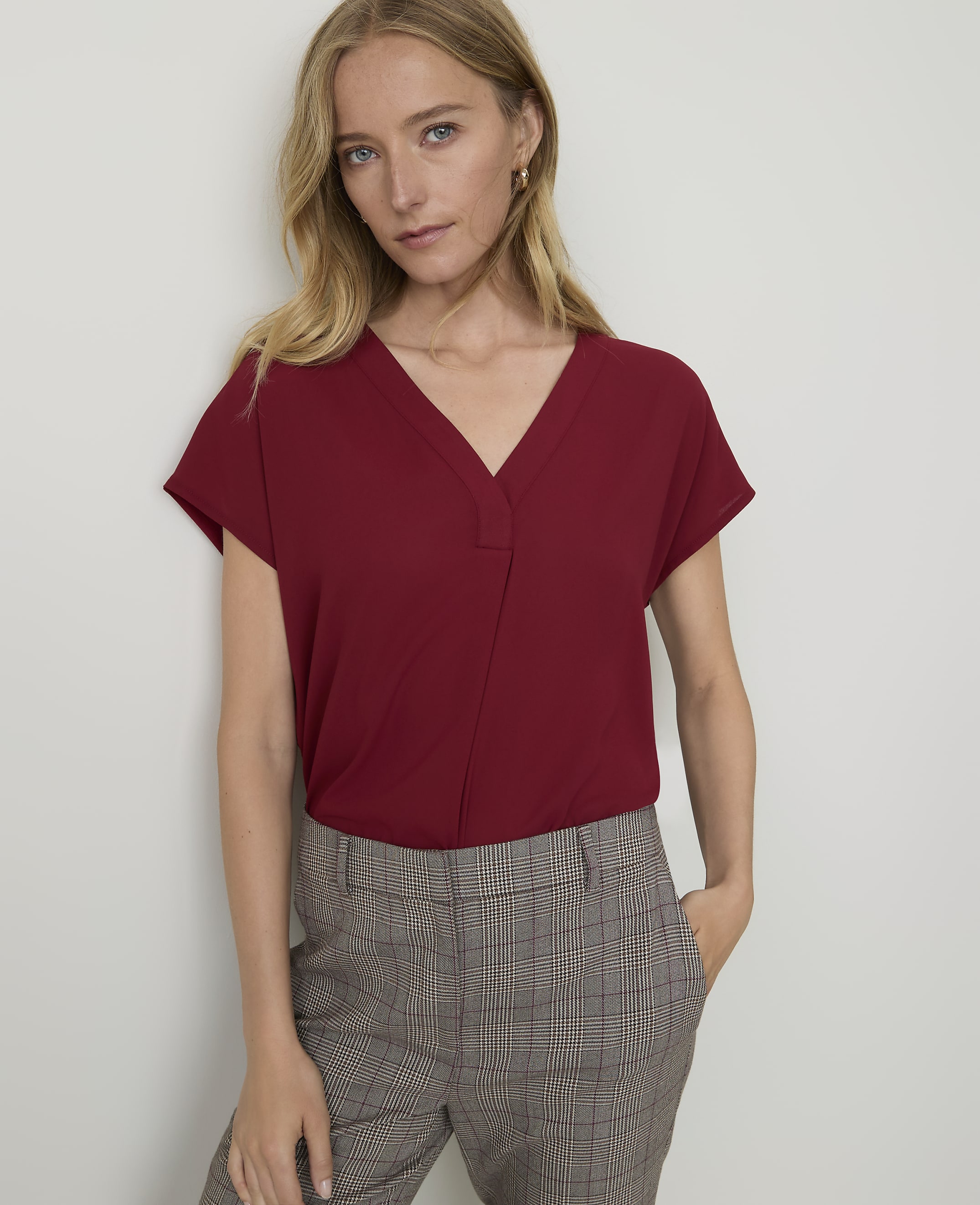 A woman in a maroon v-neck top and plaid pants stands against a plain white wall.