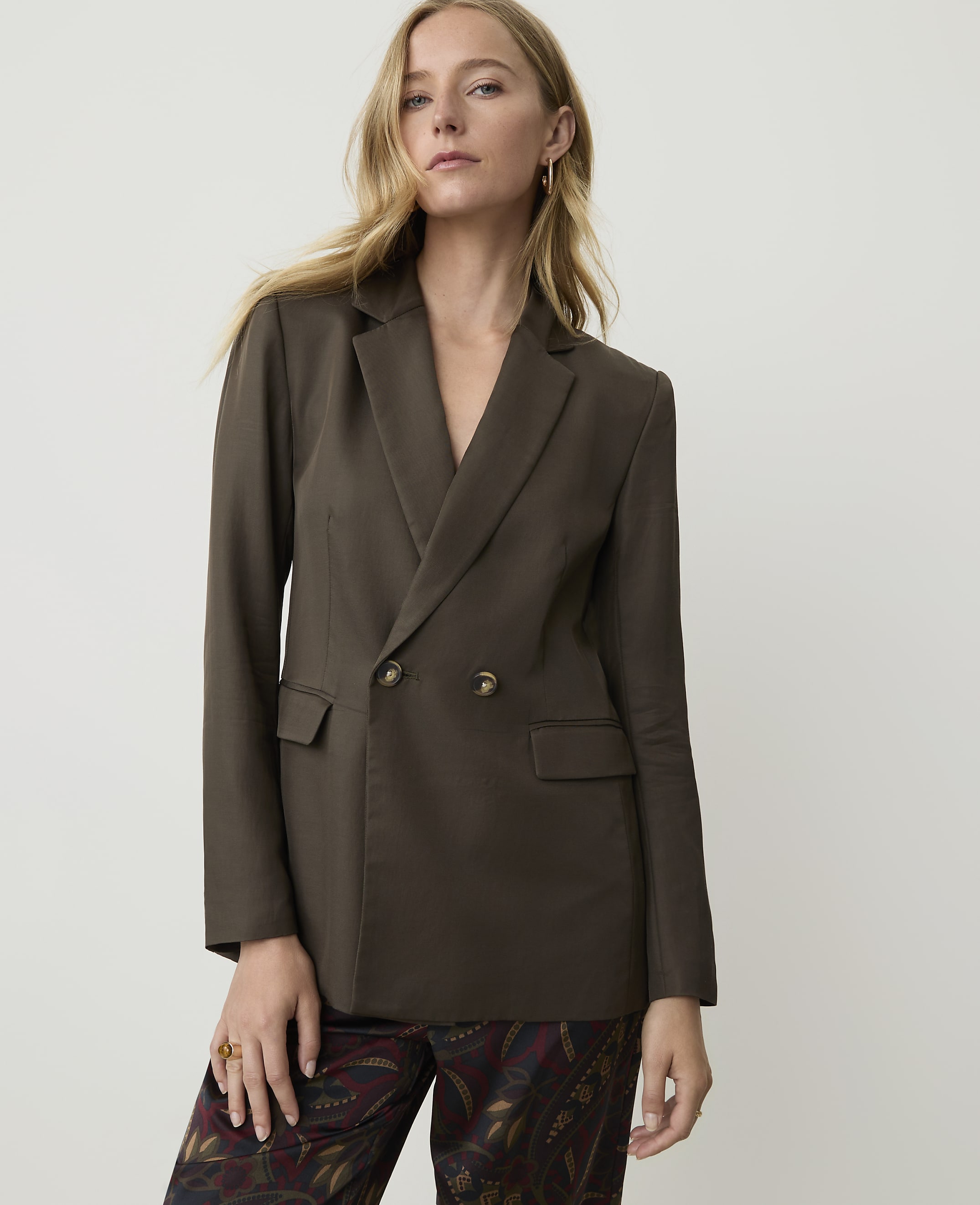 A woman in a dark brown double-breasted blazer and patterned pants stands against a plain white background.