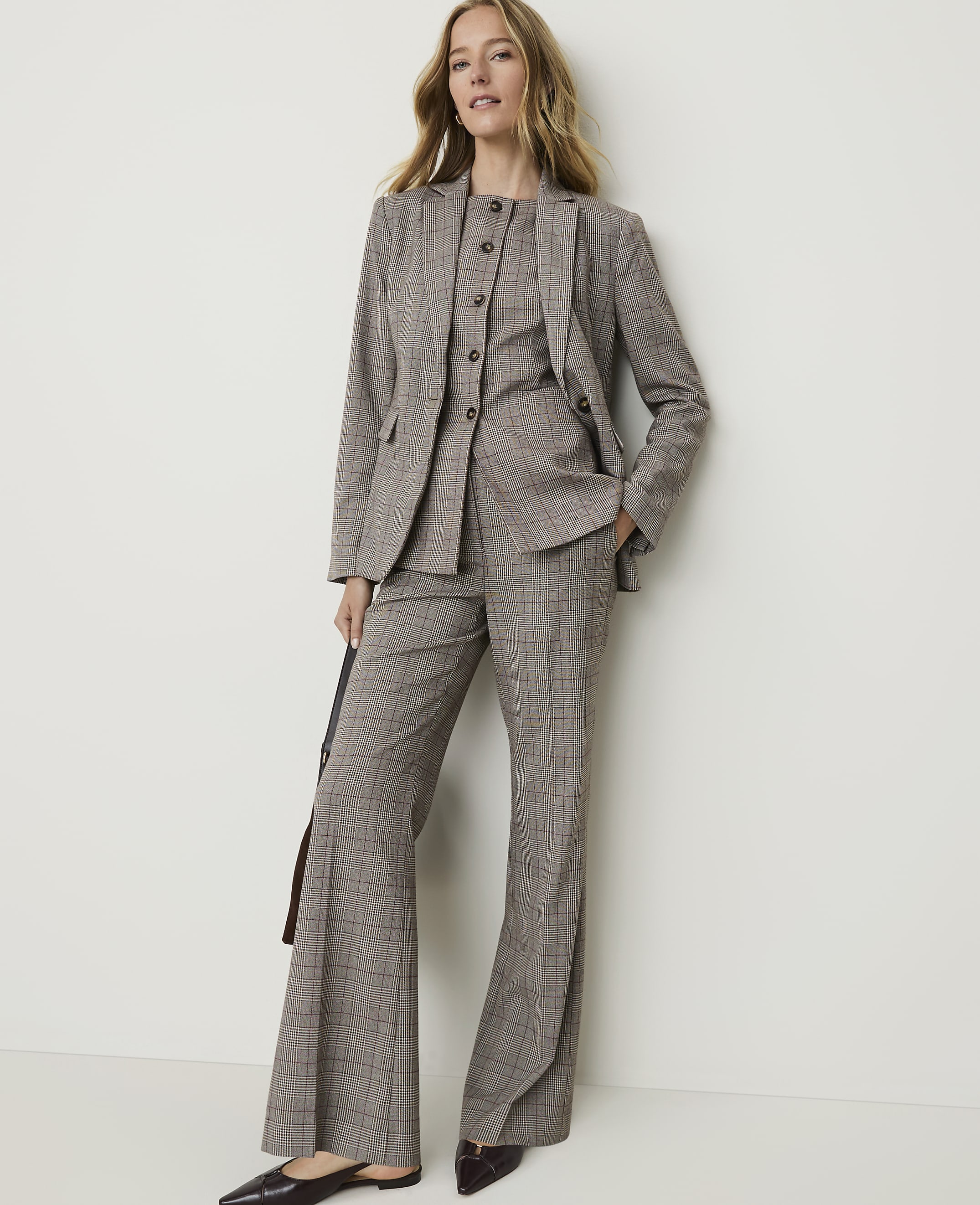 A woman in a brown plaid suit stands against a plain white wall, holding a dark bag.