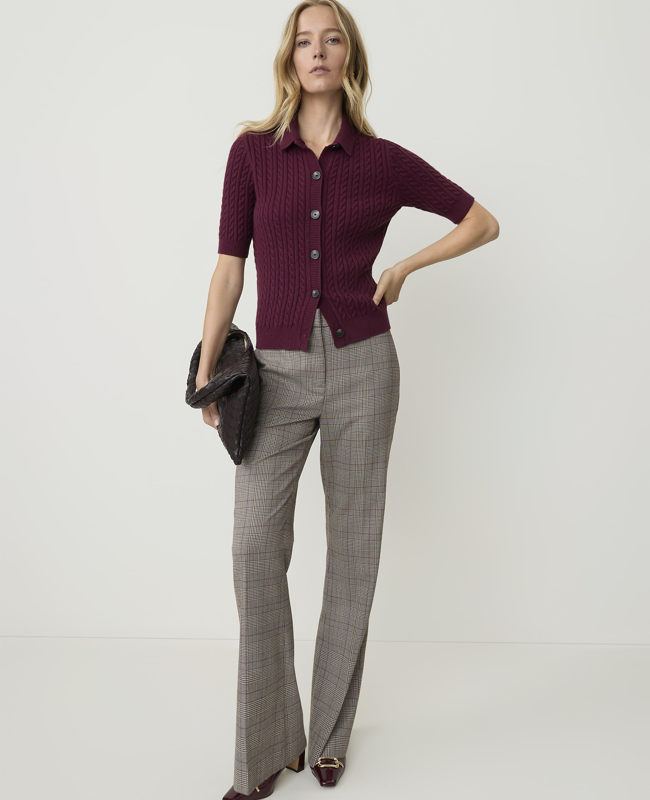 A woman in a burgundy ribbed cardigan and plaid flared pants stands against a plain white background, holding a dark clutch.