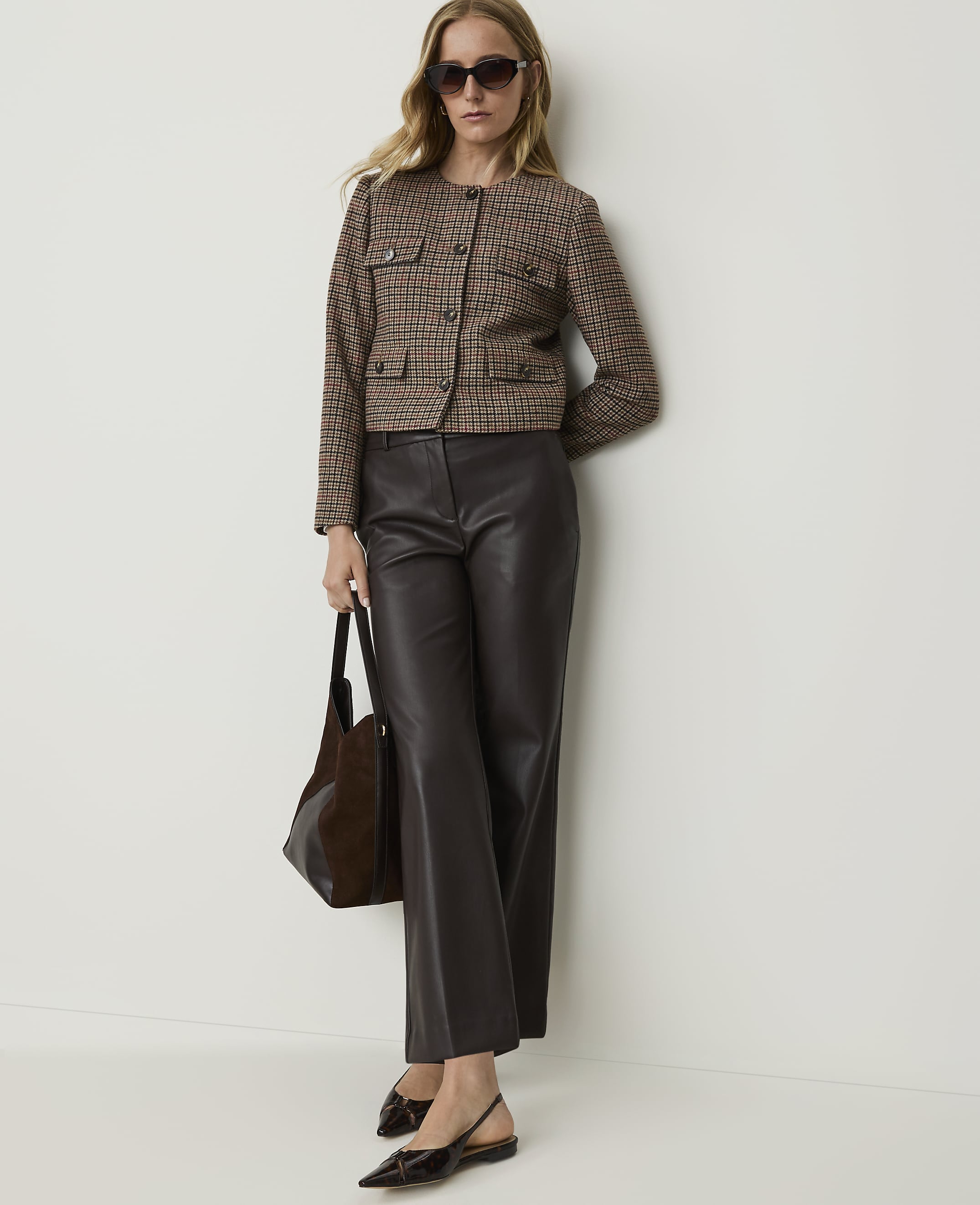 A woman in a tweed jacket and leather pants stands against a plain wall, holding a dark handbag.