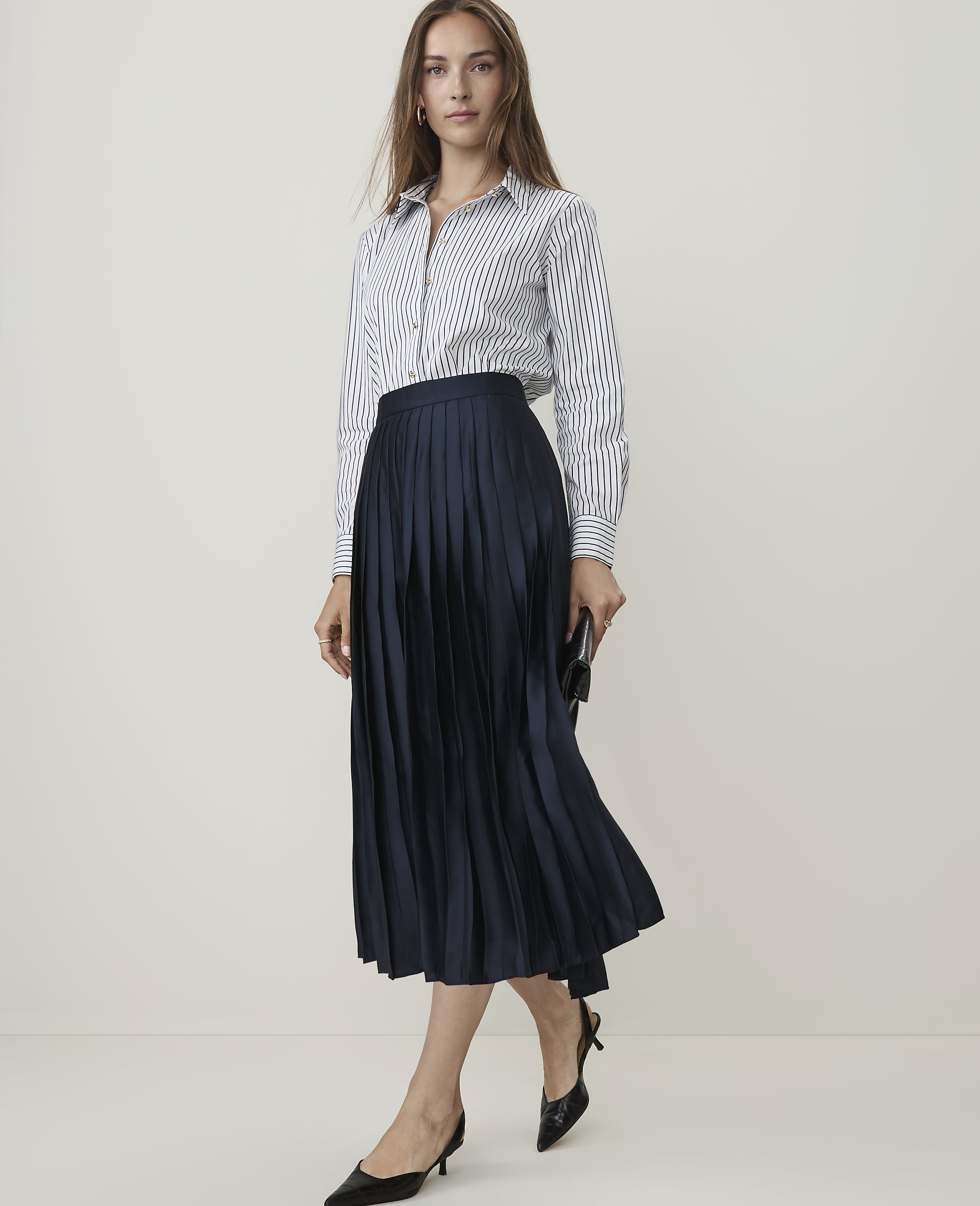 A woman in a striped shirt and navy pleated skirt stands against a plain, light background.