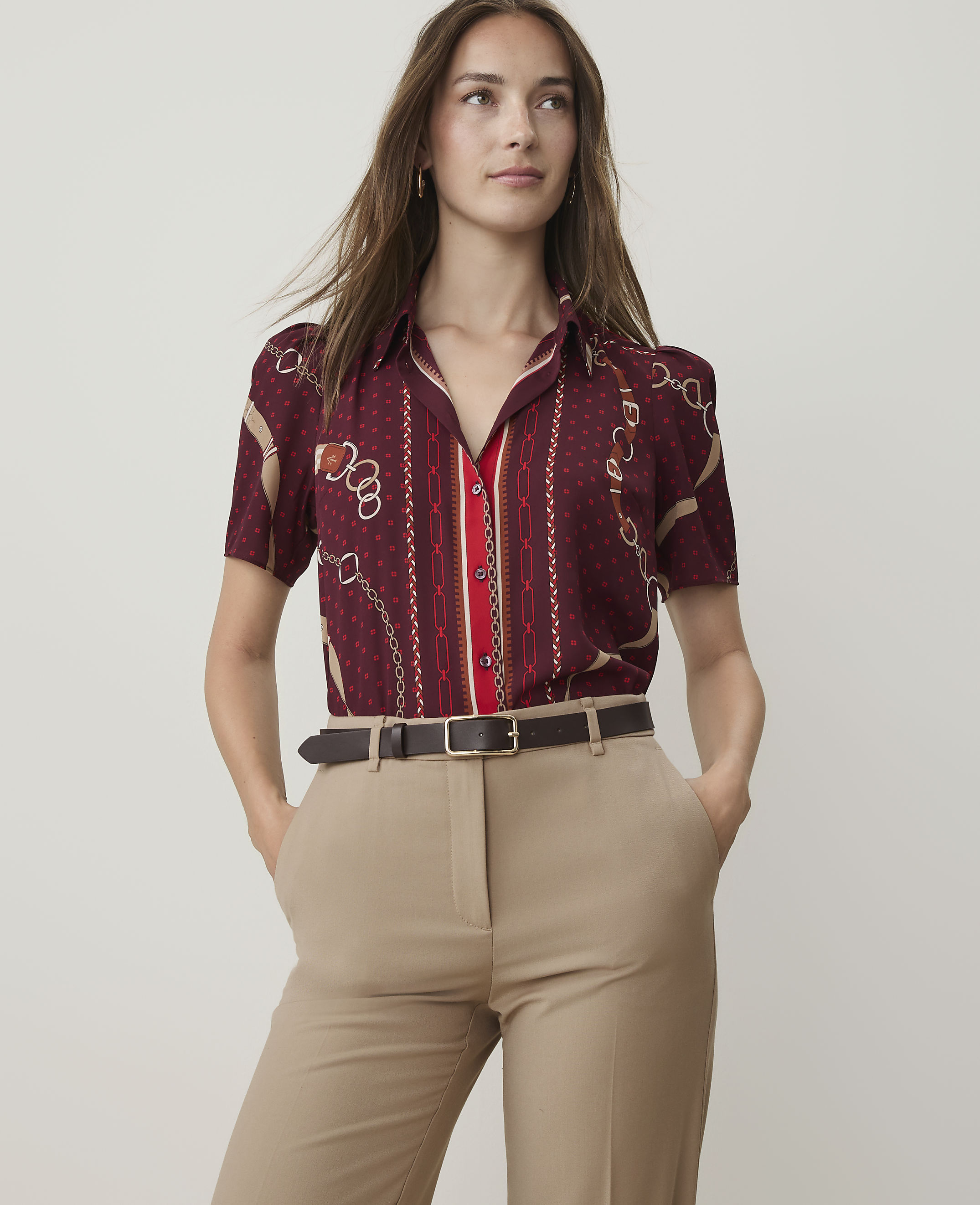 A woman in a patterned burgundy shirt and tan pants stands against a plain, light background.