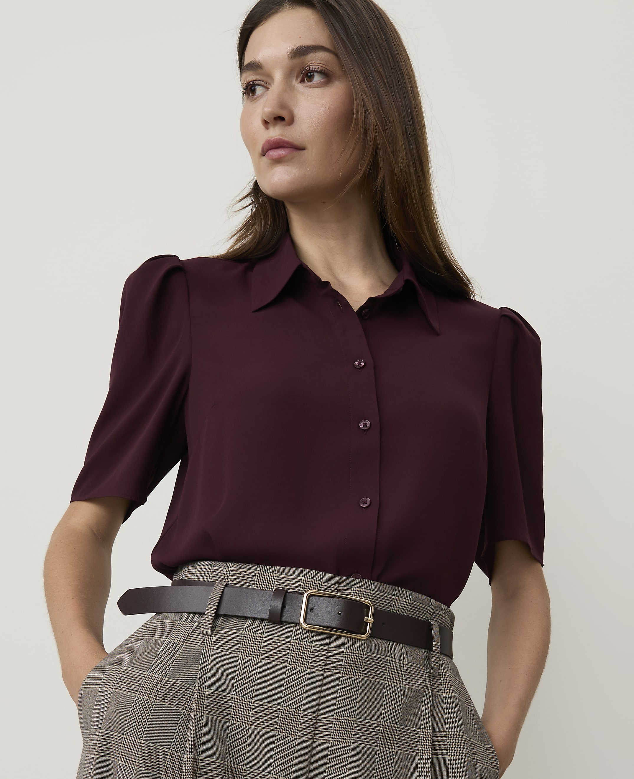 A woman in a burgundy blouse and plaid pants stands against a plain background.