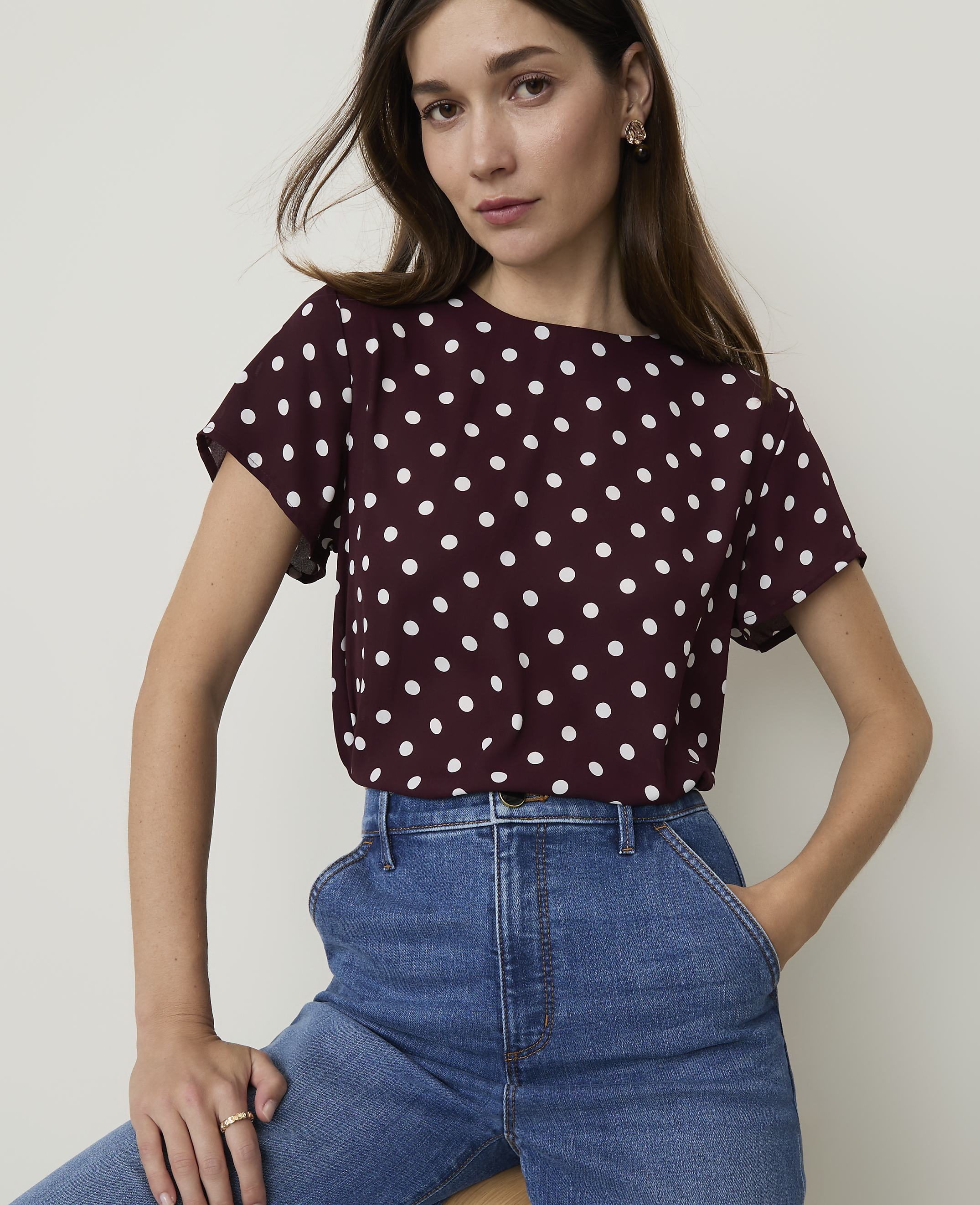 A woman in a burgundy polka dot top and blue jeans poses against a plain white background.