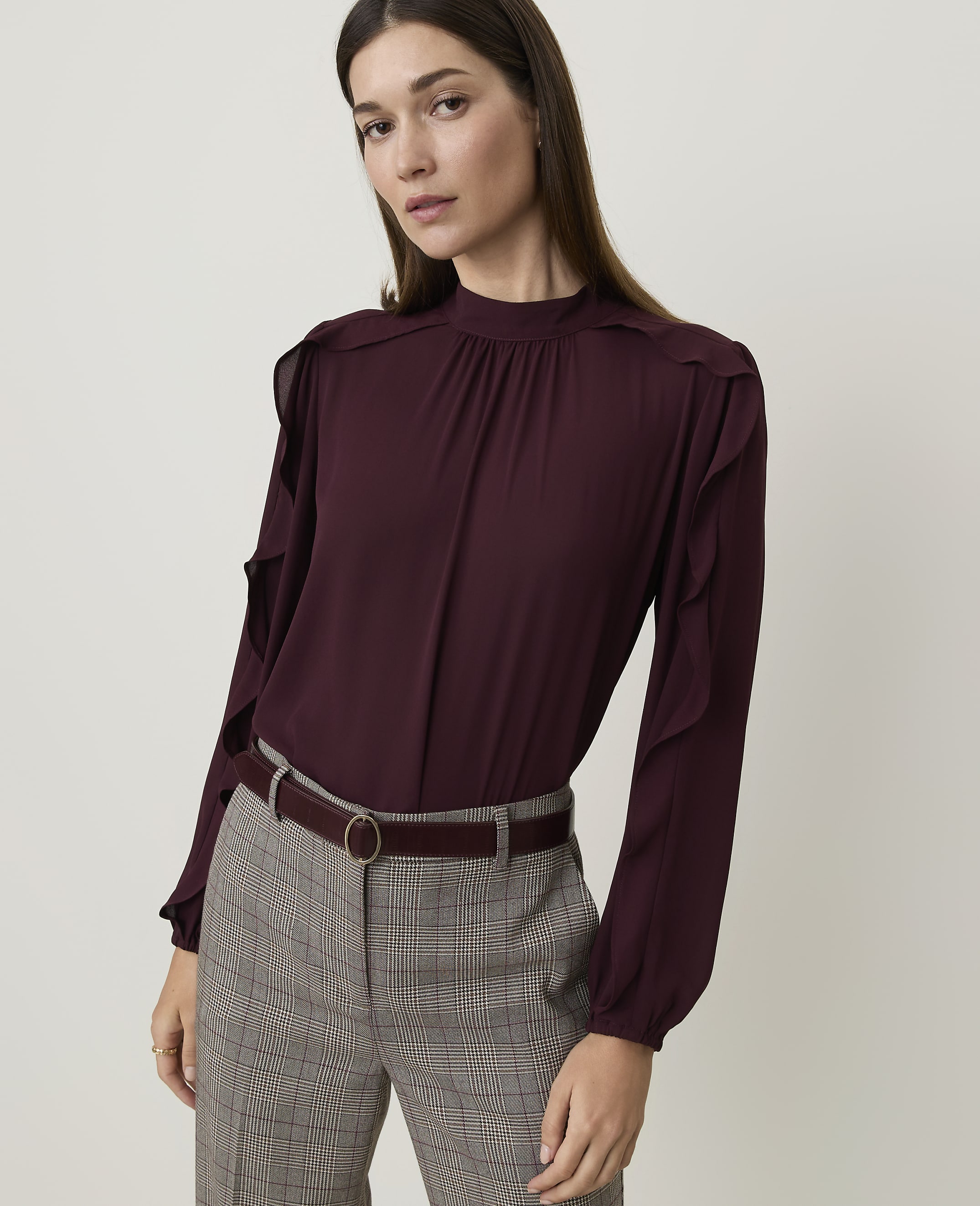 A woman in a burgundy ruffled blouse and plaid pants stands against a plain white background.