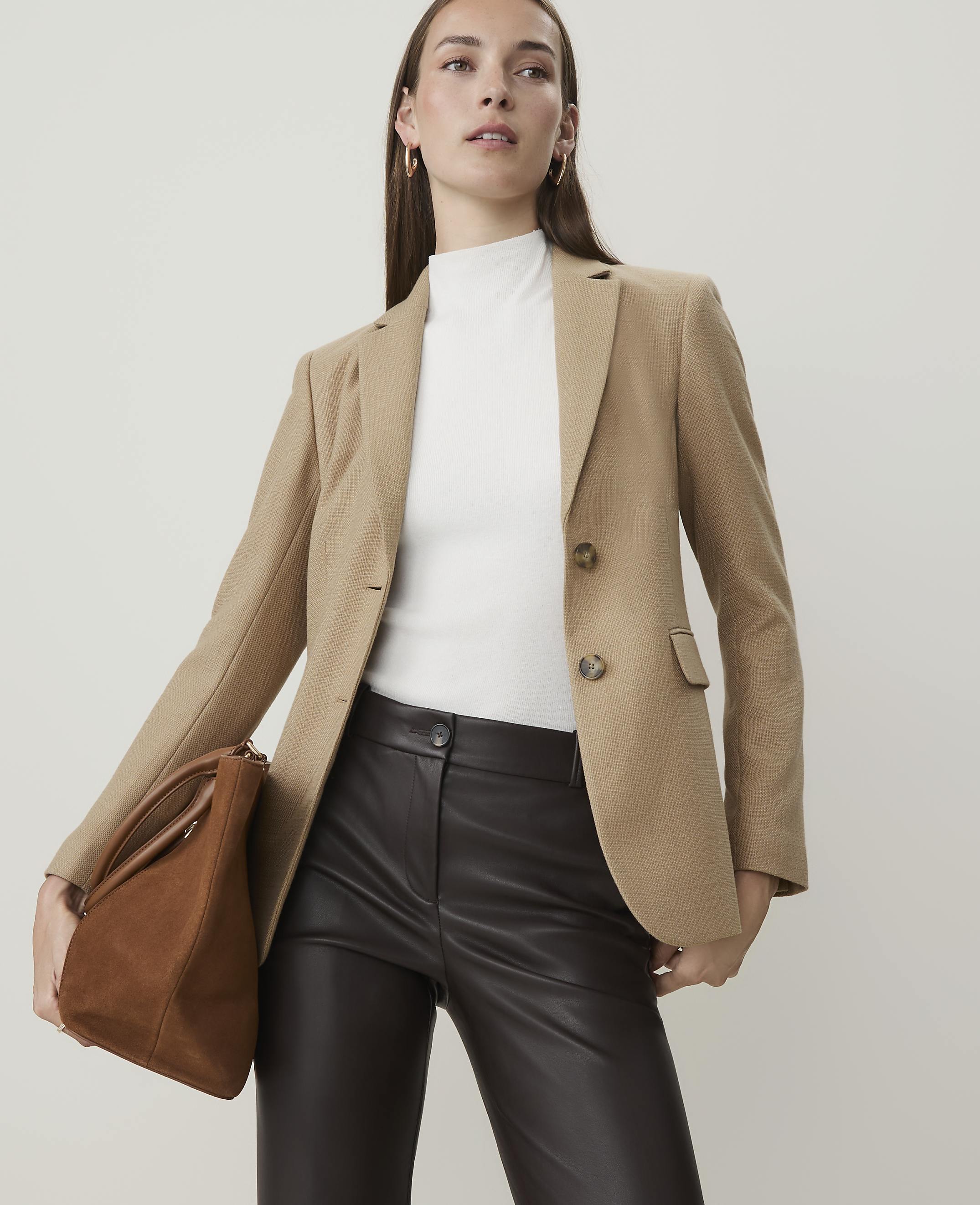A woman in a tan blazer and dark pants holds a brown purse against a plain background.