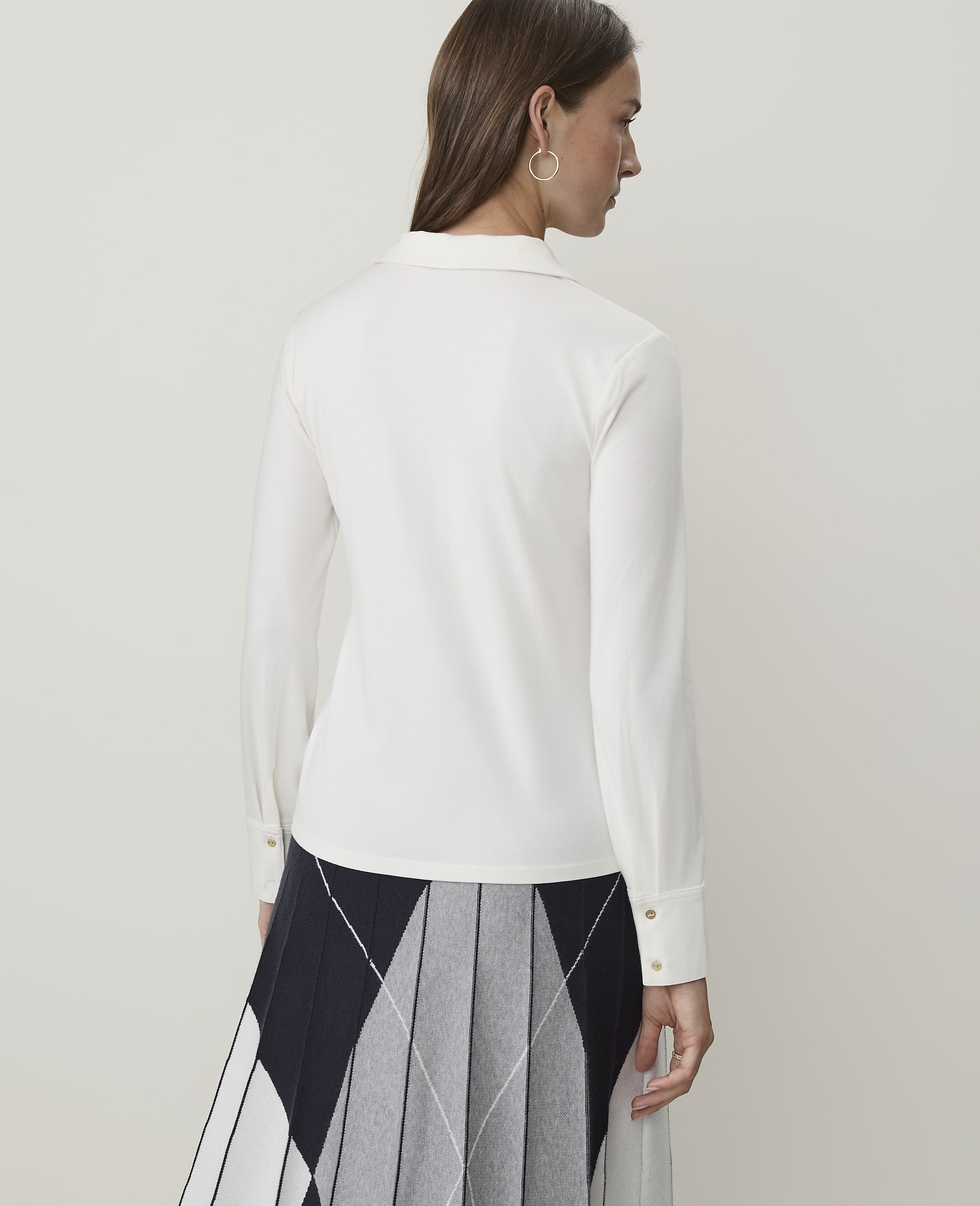 A woman in a white collared shirt and a pleated skirt stands against a plain white background.