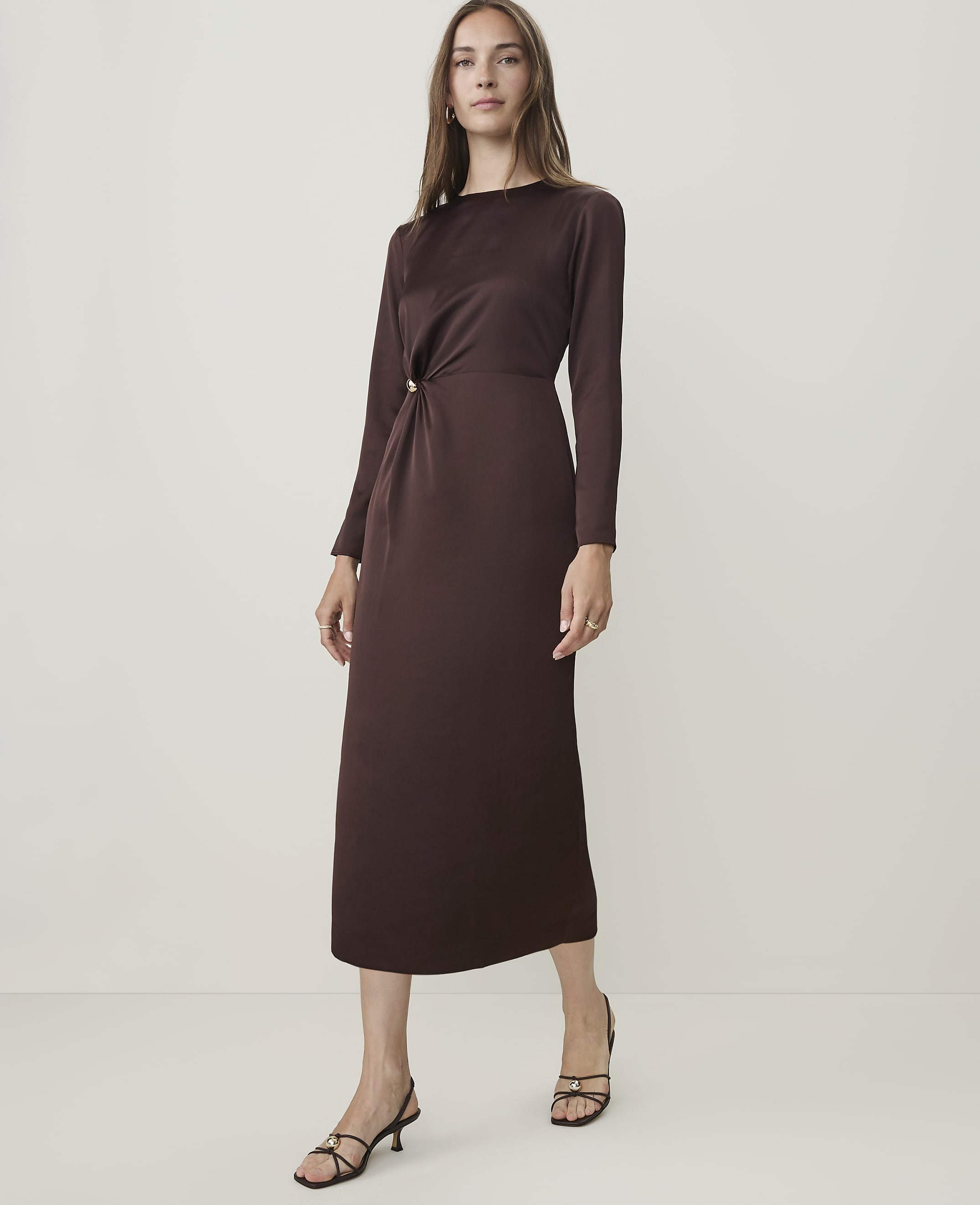 A woman in a long-sleeved brown dress stands against a plain, light-colored background.