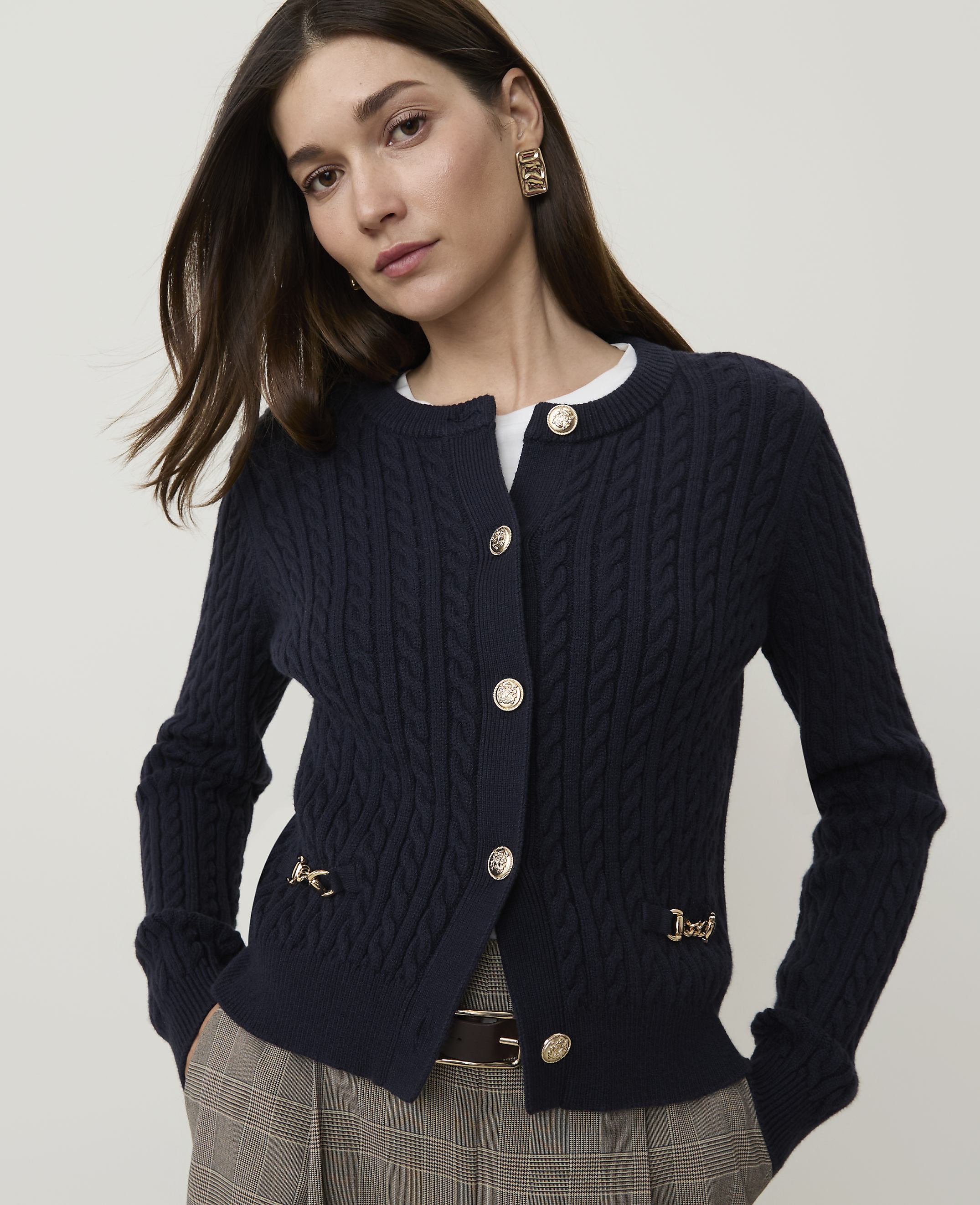 A woman in a navy cable-knit cardigan with gold buttons and chain details stands against a plain white background.
