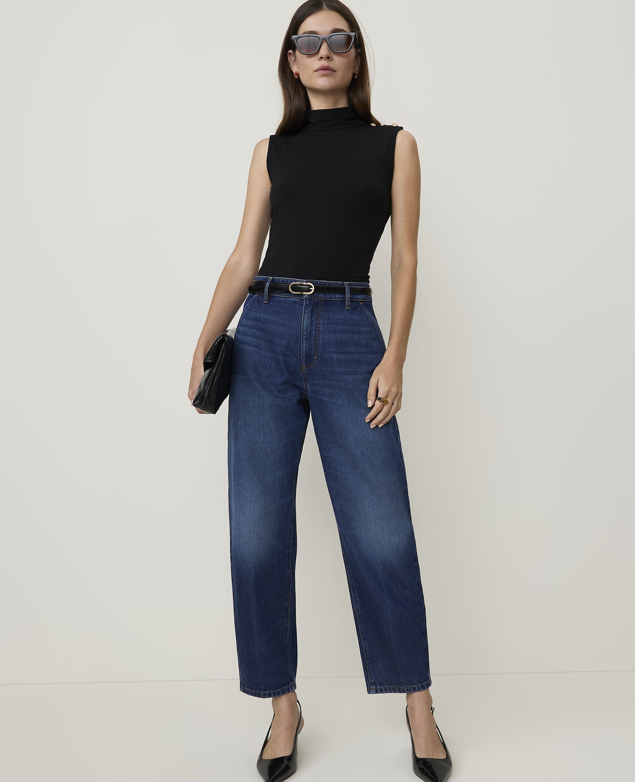 A woman in a black sleeveless top and high-waisted jeans stands against a plain white background, holding a black clutch.