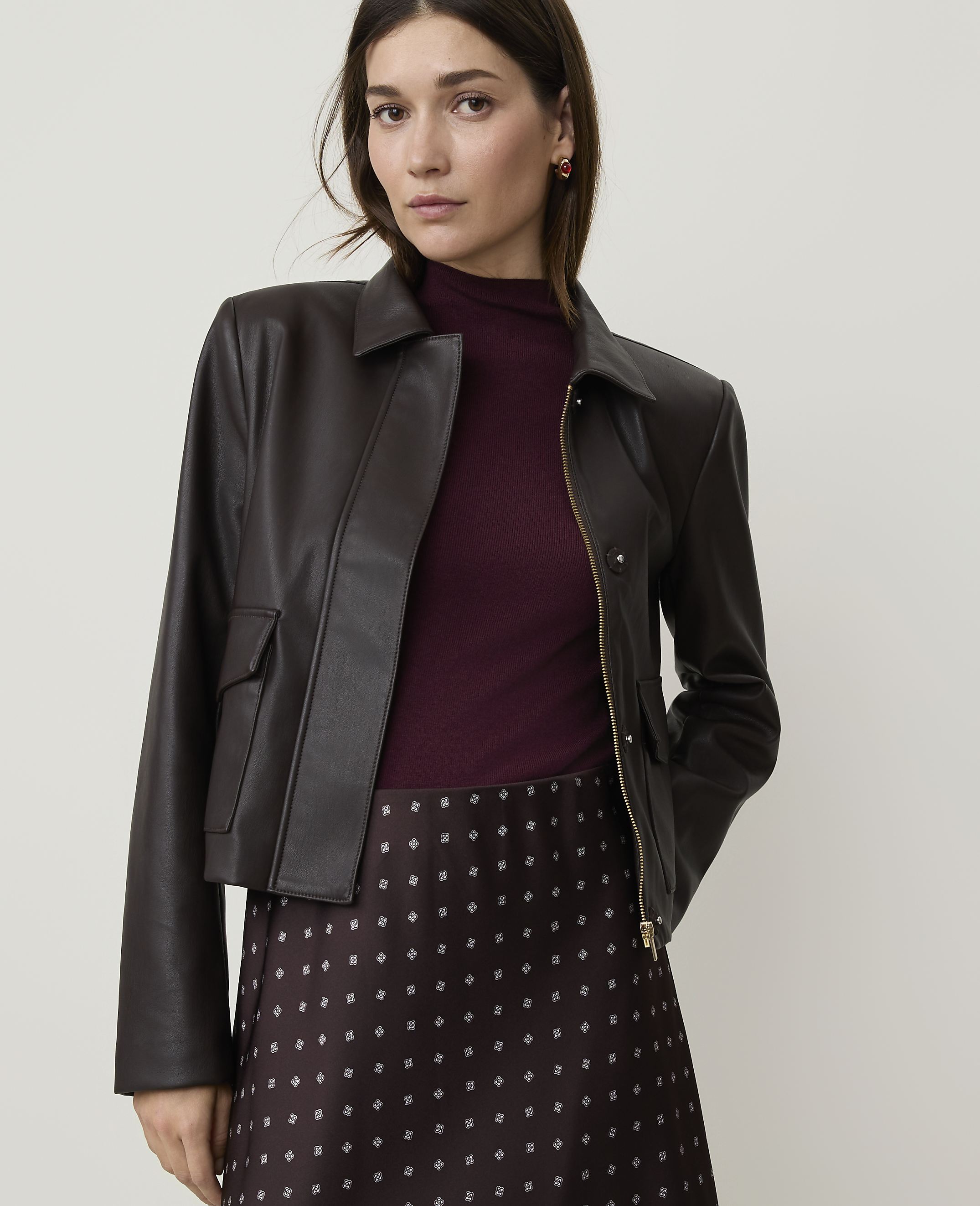 A woman in a dark brown faux leather jacket and burgundy turtleneck stands against a plain background.