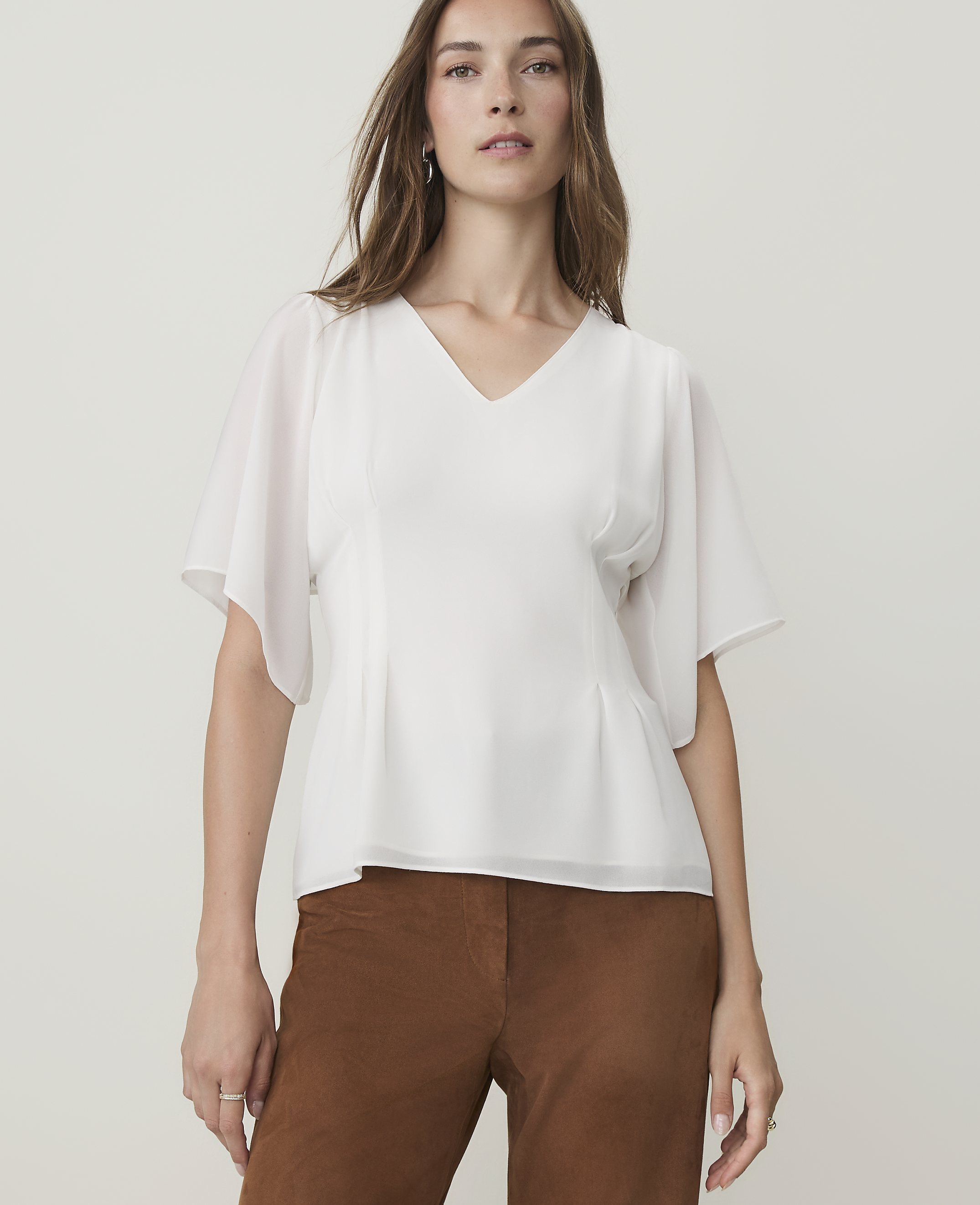 A woman in a white v-neck blouse and brown pants stands against a plain, light gray background.