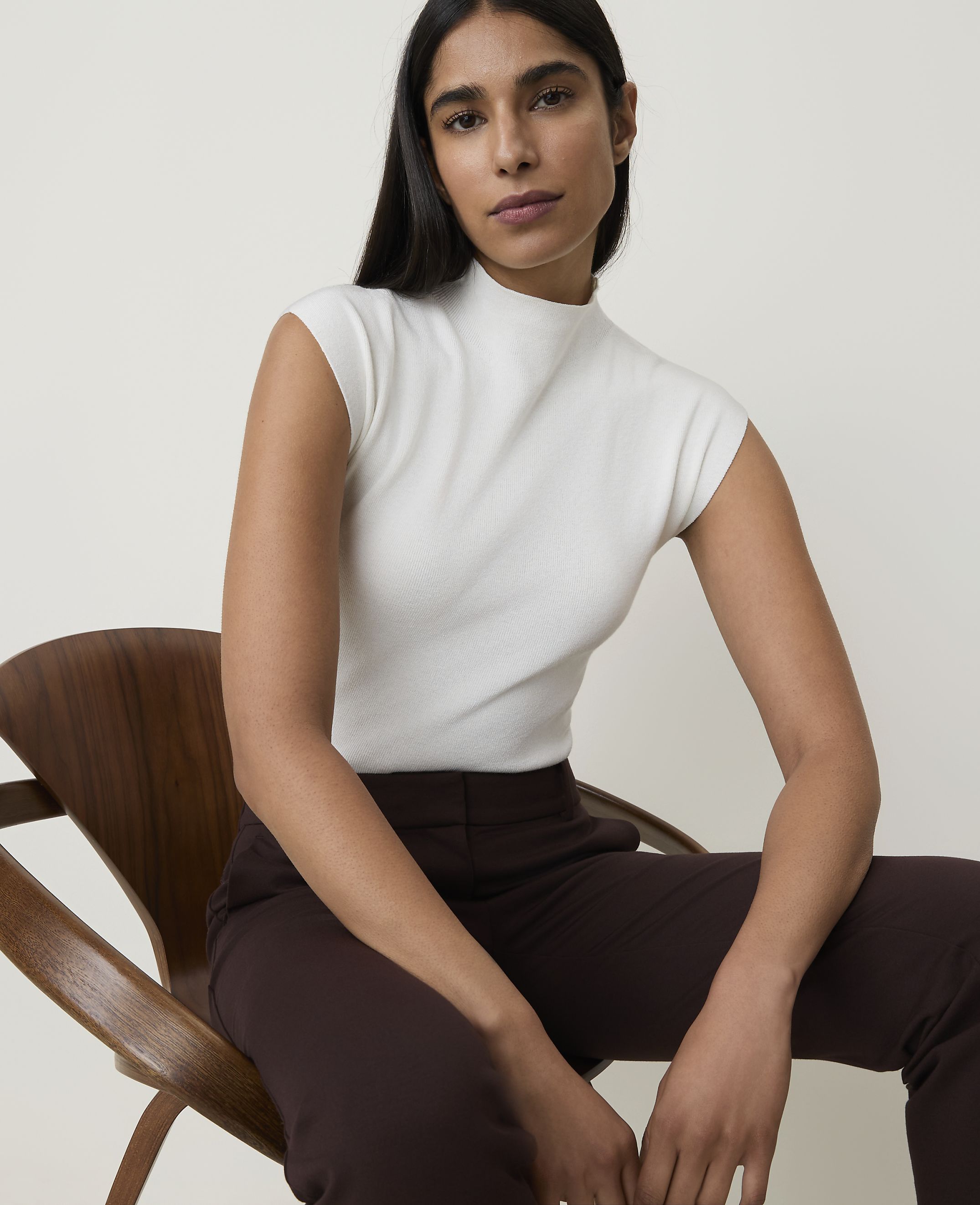A woman in a white mock-neck top and brown pants sits on a wooden chair against a plain, light background.