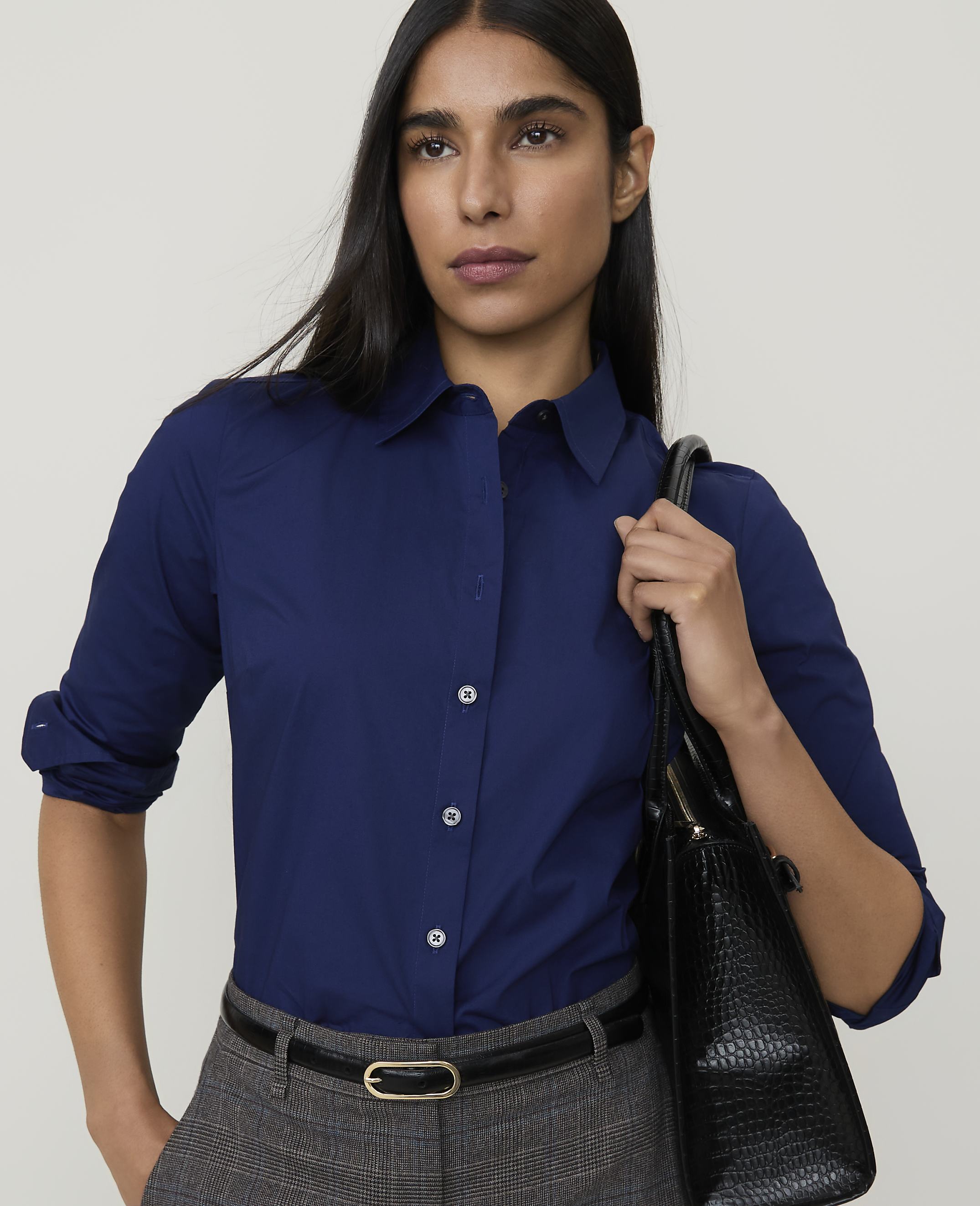 A woman in a navy blue collared shirt and grey pants, carrying a black handbag, stands against a plain grey background.