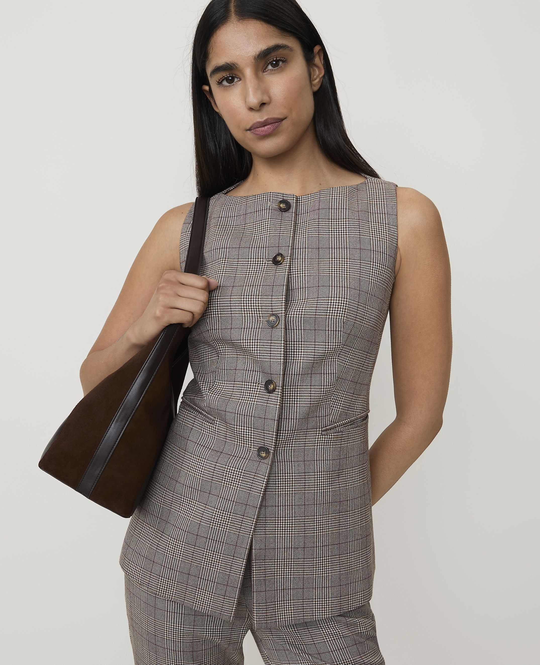 A woman in a plaid vest and pants carries a brown bag against a plain background.
